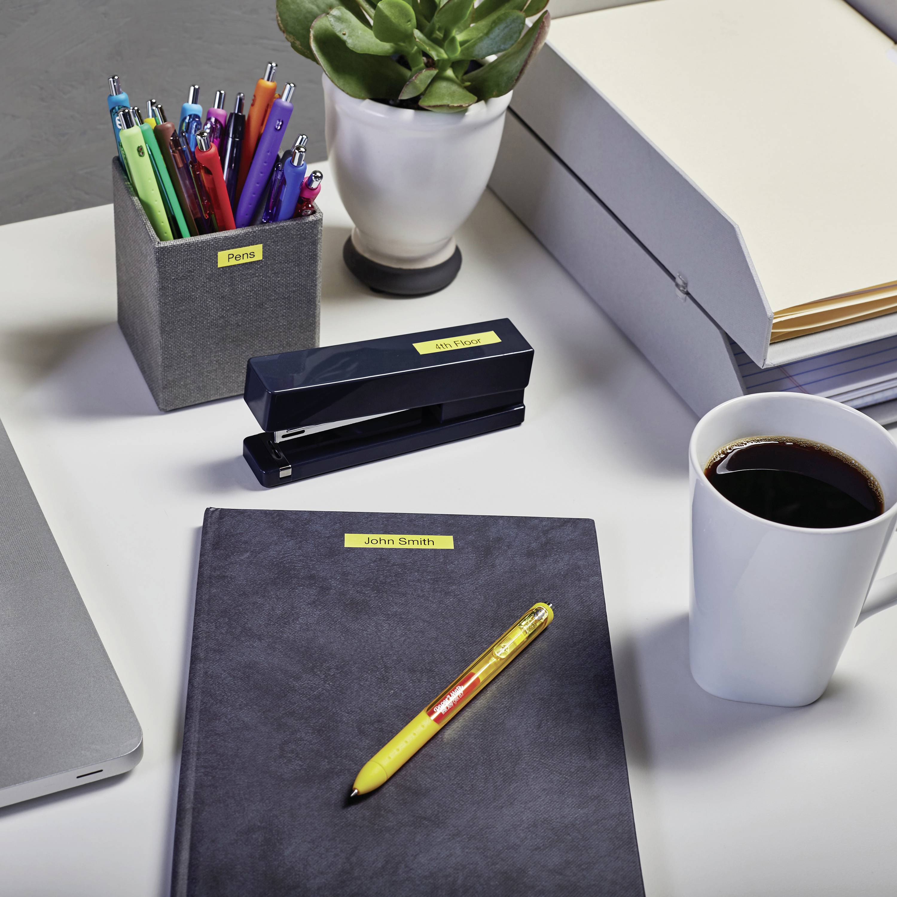 Schreibtisch mit Laptop, Stiften, Heftklammerhalter, Pflanze, Ordnerstapel, Notizbuch und einer Tasse Kaffee.