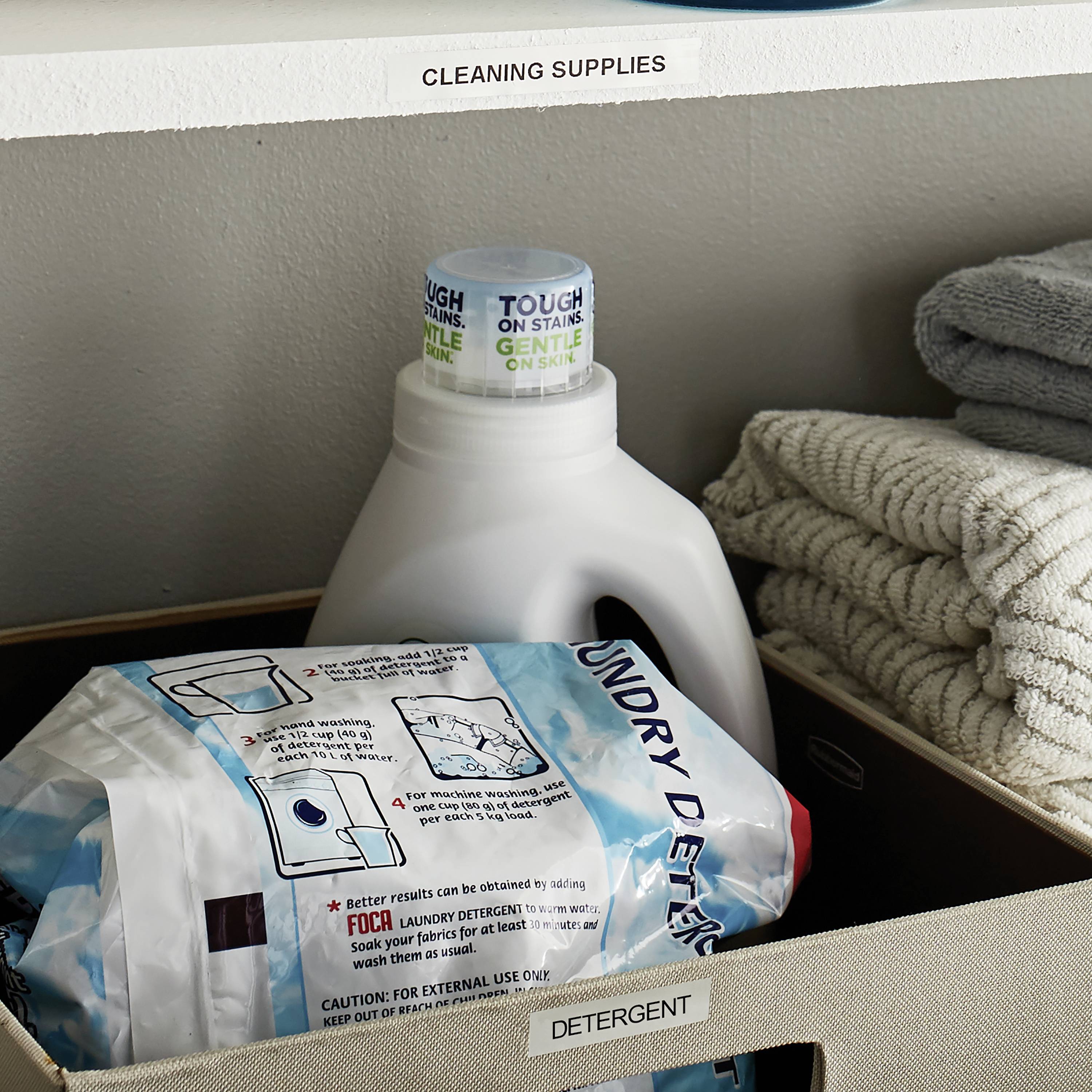 Zwei Flaschen Waschmittel und ein Karton mit gefalteten Handtüchern auf einem Regal mit der Beschriftung 'Cleaning Supplies'.