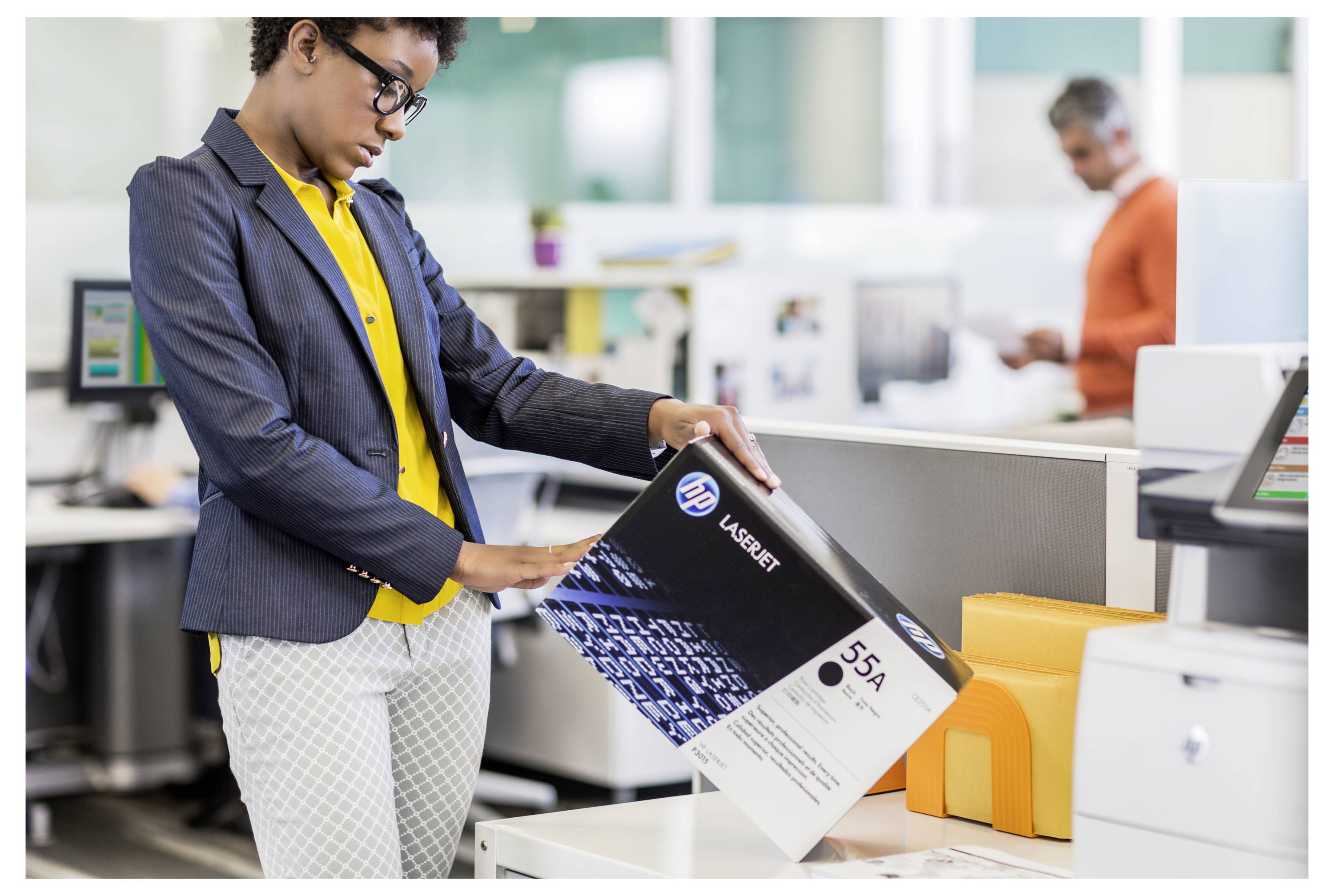 Eine Frau in einem Büro wählt eine LaserJet 55A-Tonerbox aus einem Stapel auf dem Schreibtisch aus, während ein Kollege im Hintergrund arbeitet.