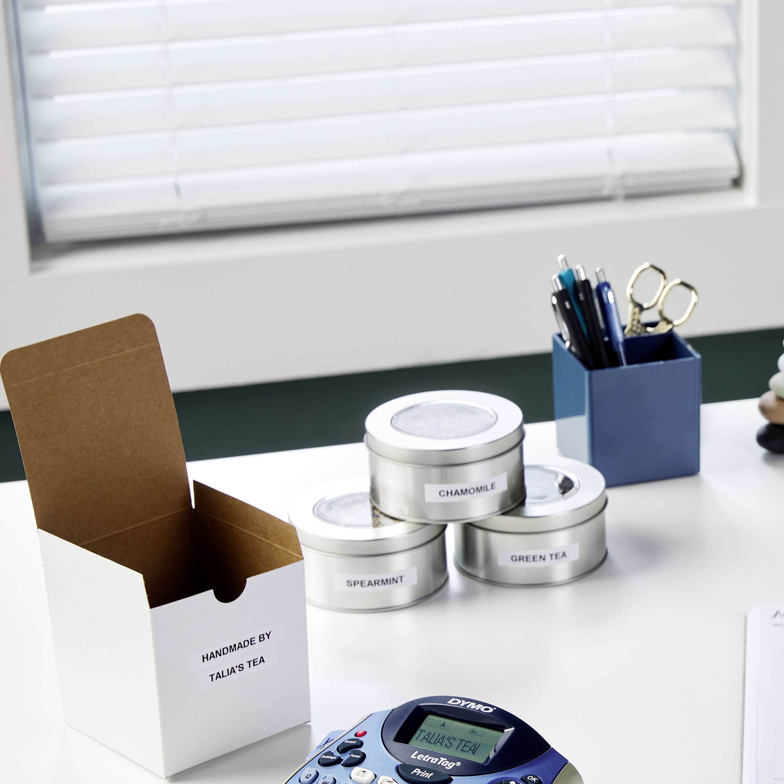 Schreibtisch mit Teedosen 'Lavender', 'Chamomile', 'Green Tea', Stiften in einem Halter, einer Teeschachtel von 'Talias Tea' und einem Beschriftungsgerät.