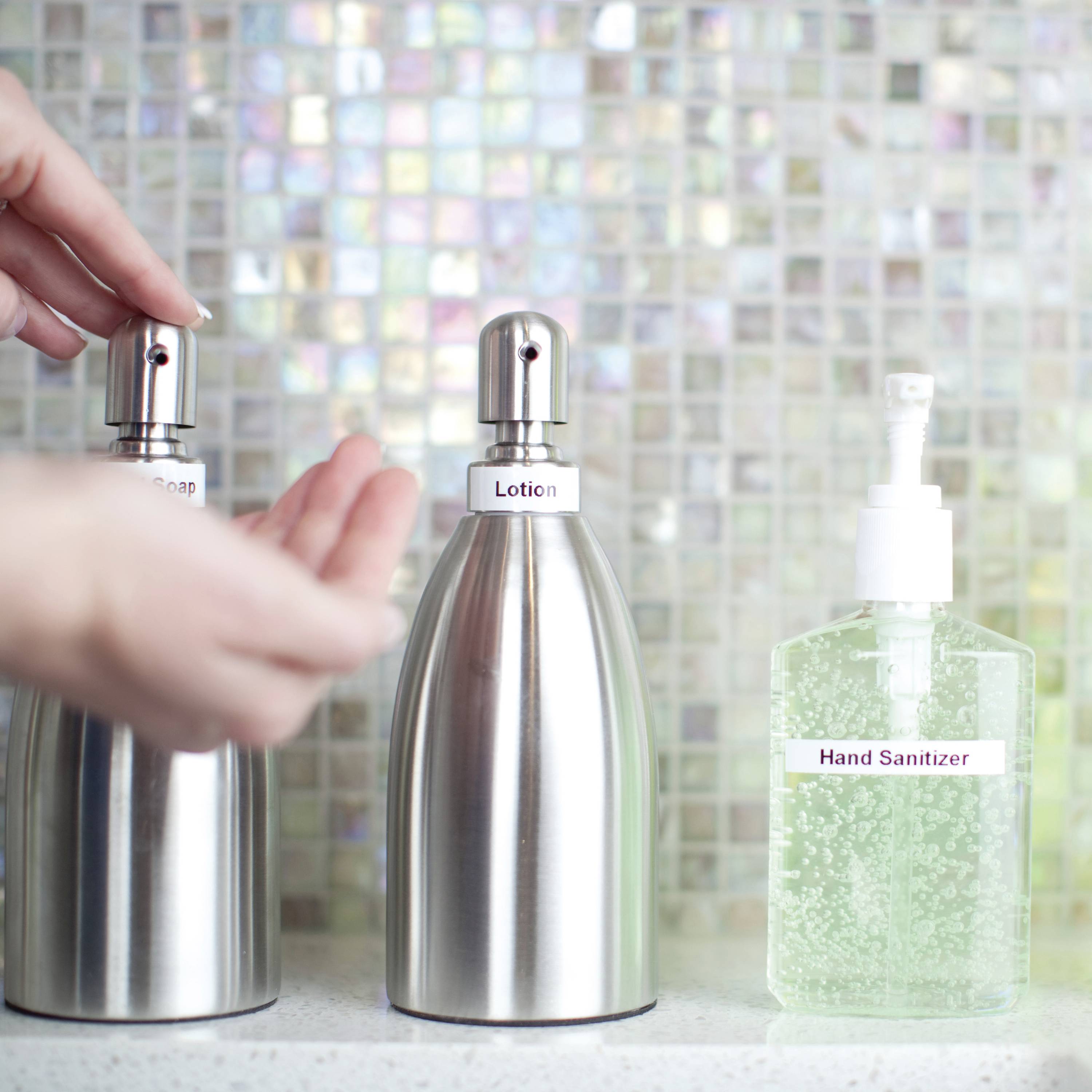 Person pumpt Seife aus einem Spender vor einem Mosaikhintergrund. Daneben stehen ein Lotion- und ein Handdesinfektionsmittelspender.