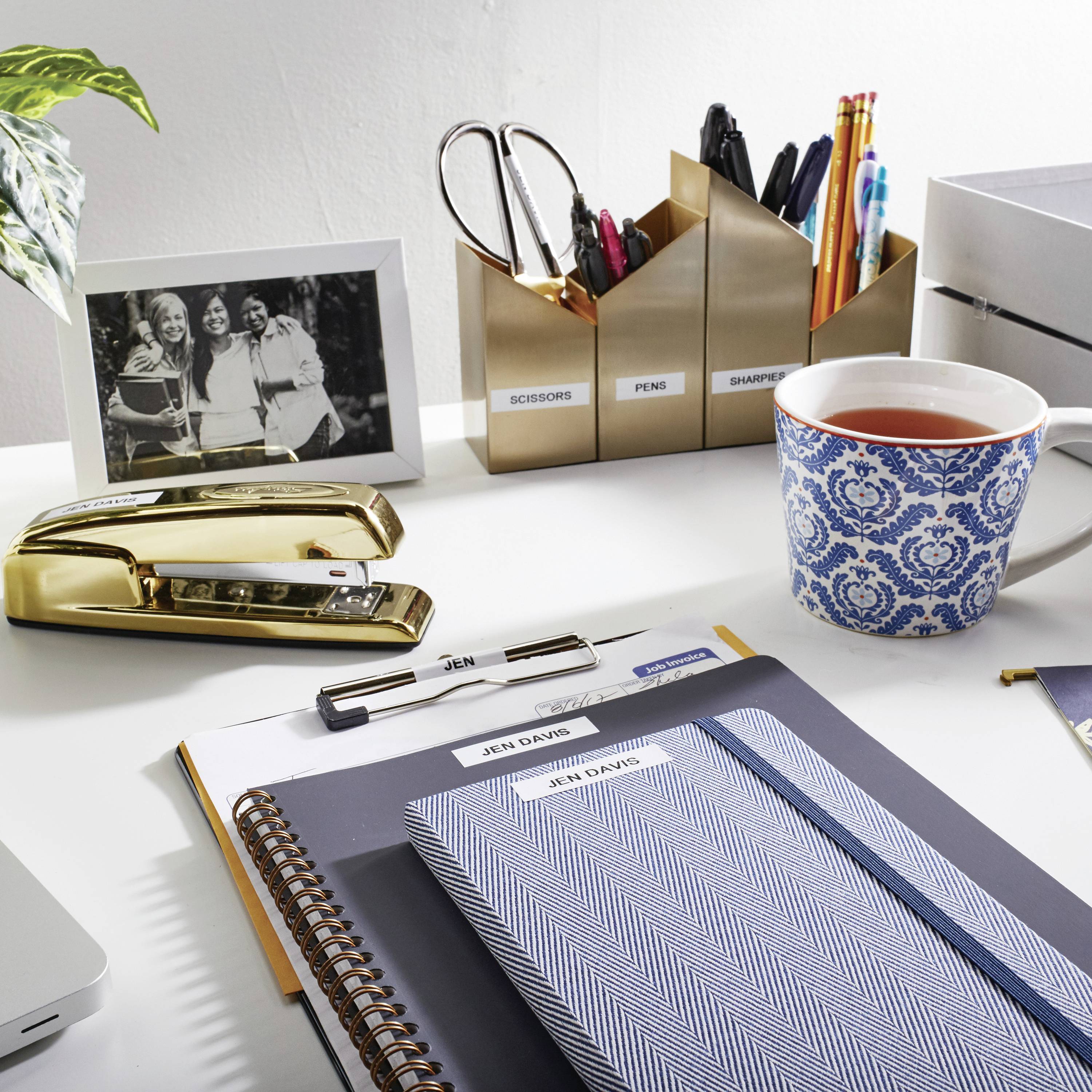 Ein organisierter Schreibtisch mit Tasse Tee, Stiftetöpfen, Notizbuch und gerahmtem Schwarz-Weiß-Foto von drei lachenden Personen.