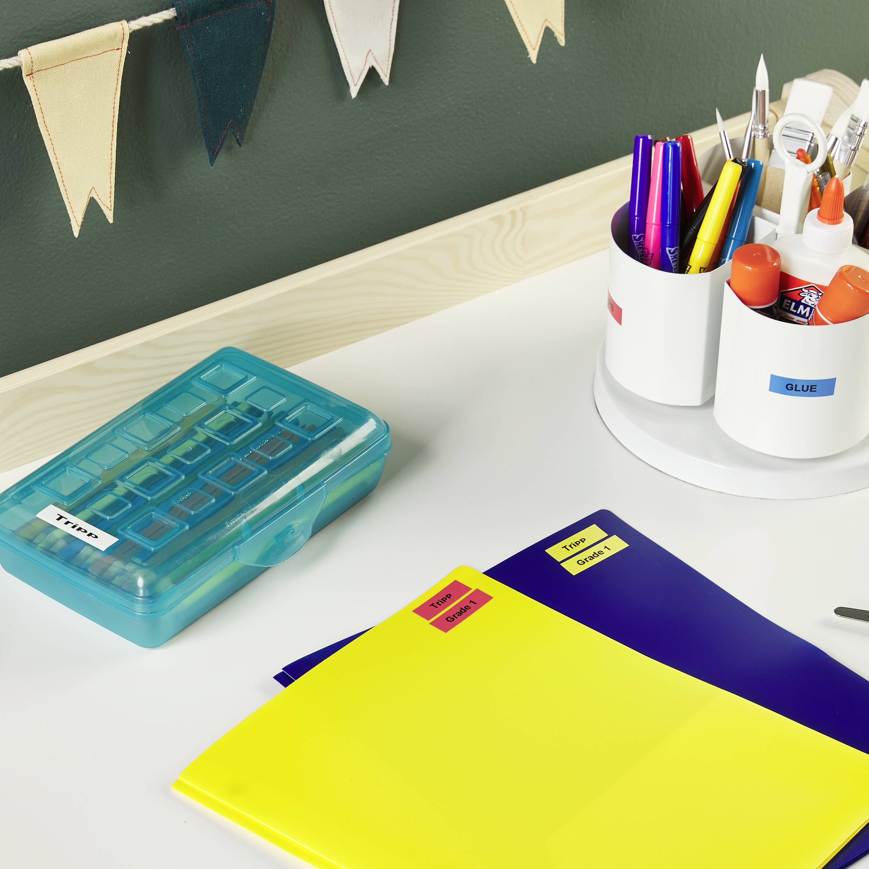 Bürobedarf auf einem Schreibtisch: blaue und gelbe Ordner, farbige Stifte in einem Behälter, eine Box mit Etiketten und Wimpel an der Wand.