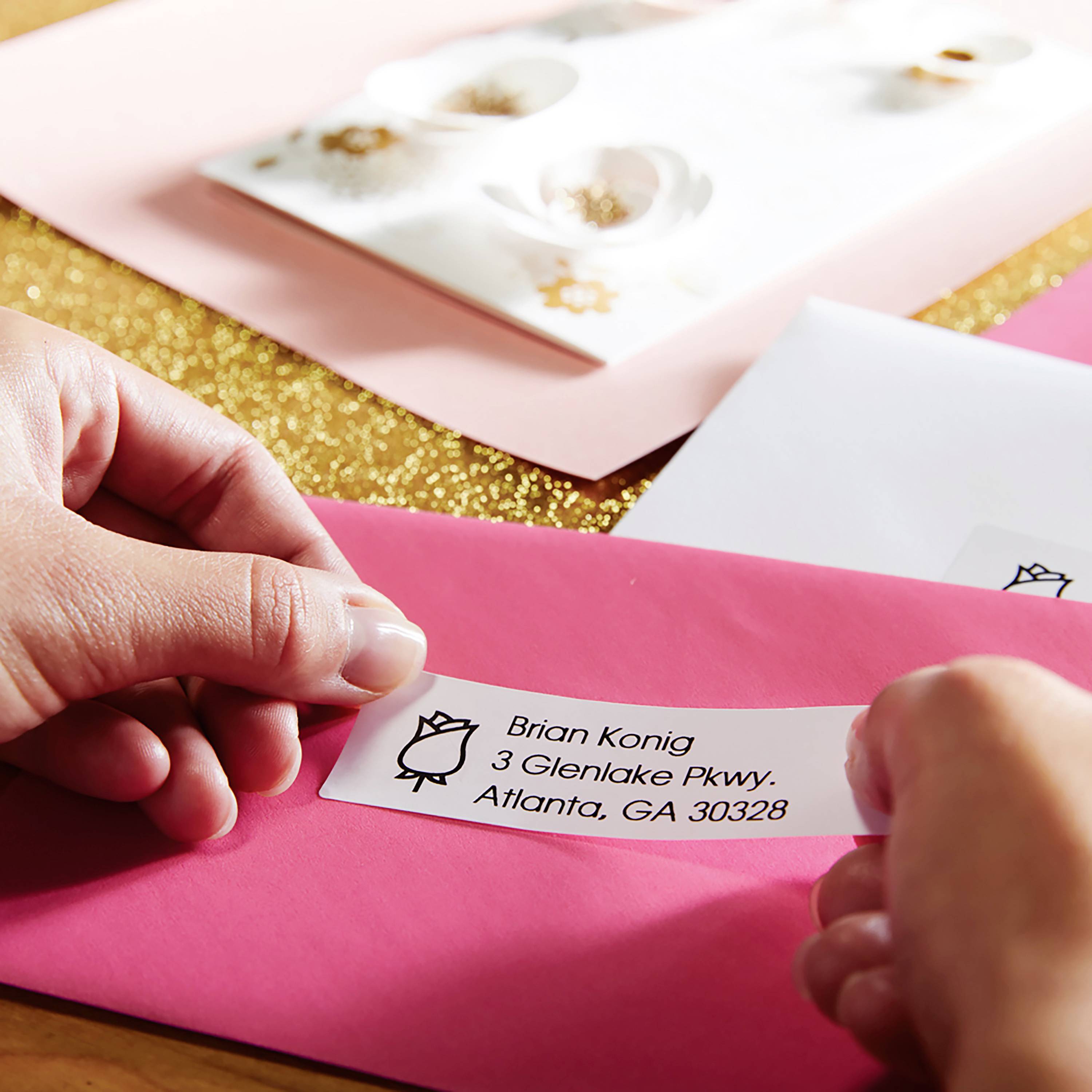 Eine Hand klebt ein Adressetikett auf einen rosa Umschlag. Der Hintergrund zeigt weitere Umschläge und eine goldene Oberfläche.