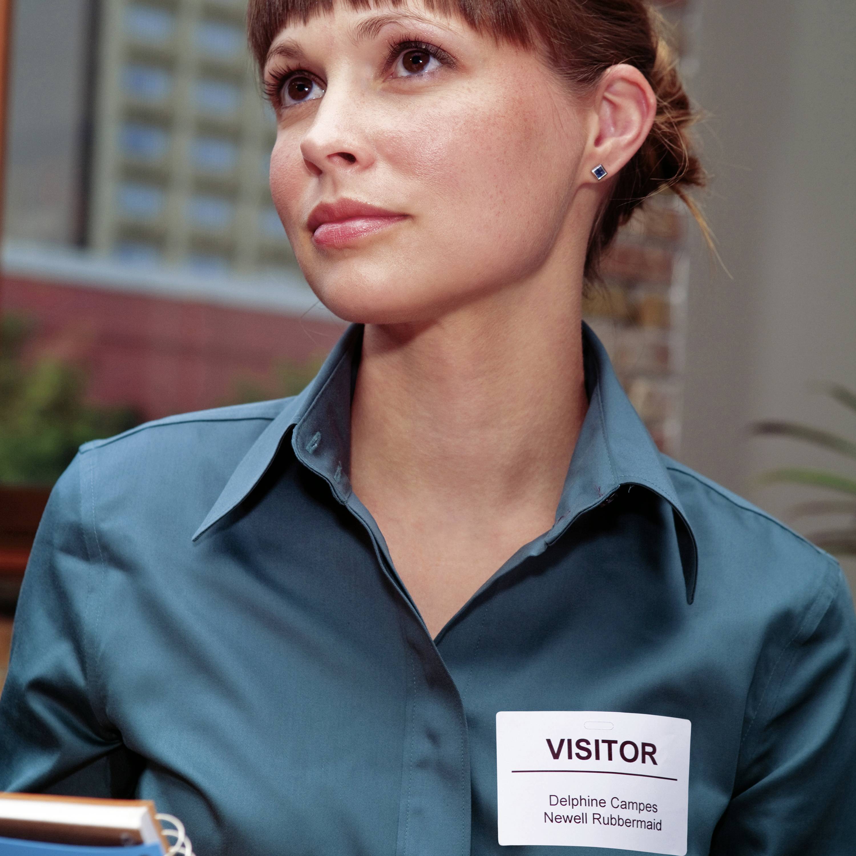 Eine Person in einem blauen Hemd trägt ein Namensschild mit der Aufschrift 'Visitor'. Sie blickt nachdenklich zur Seite.