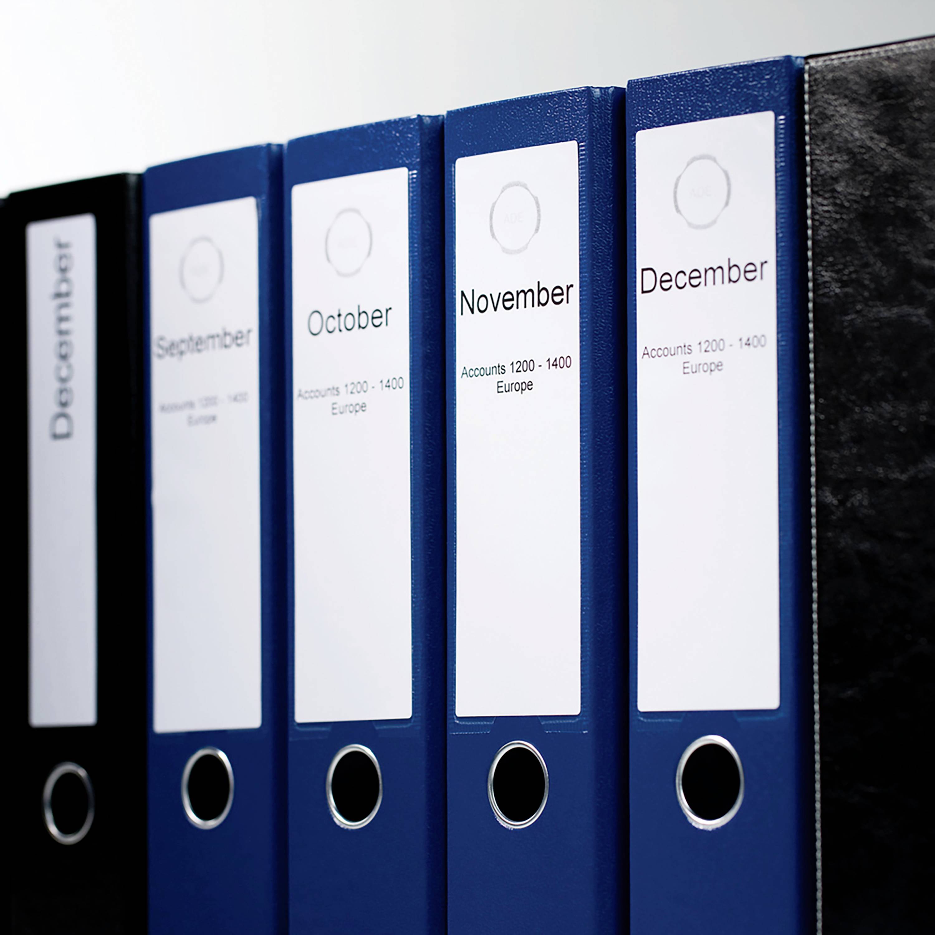 Ordnerreihe mit Beschriftungen: 'December', 'September', 'October', 'November', 'Accounts'. Steht auf einem Regal.
