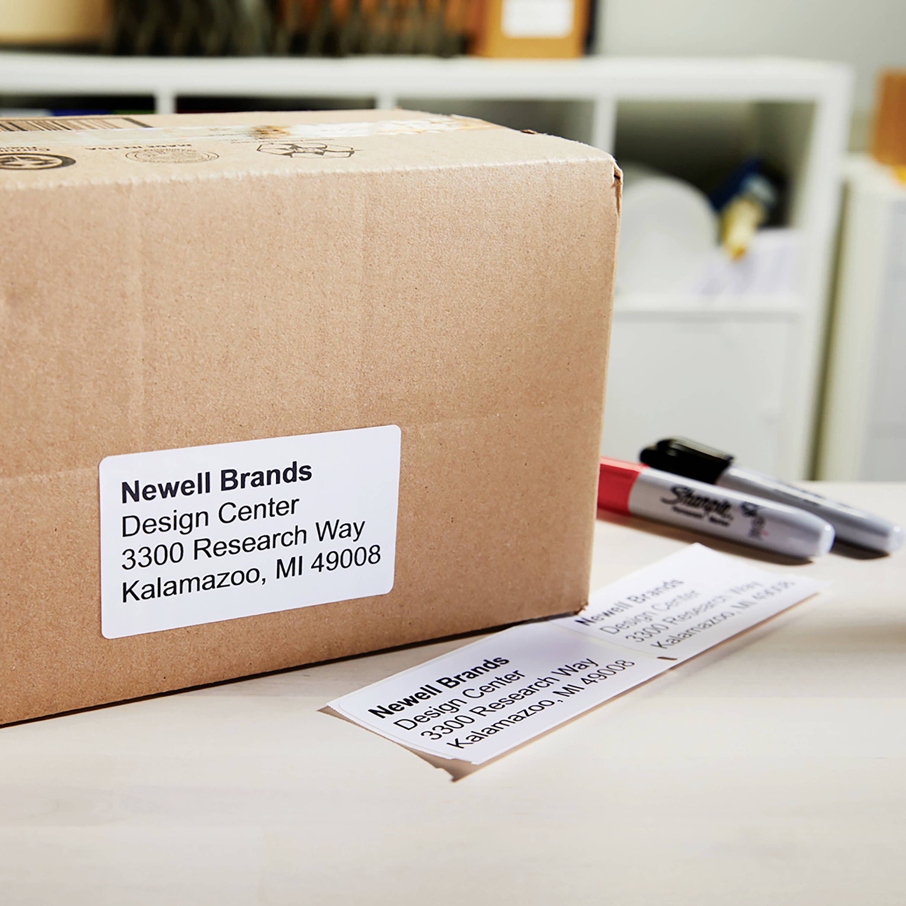 Paket mit Aufschrift 'Newell Brands, Design Center, 3300 Research Way, Kalamazoo, MI 49008' auf einem Tisch, daneben zwei Marker.