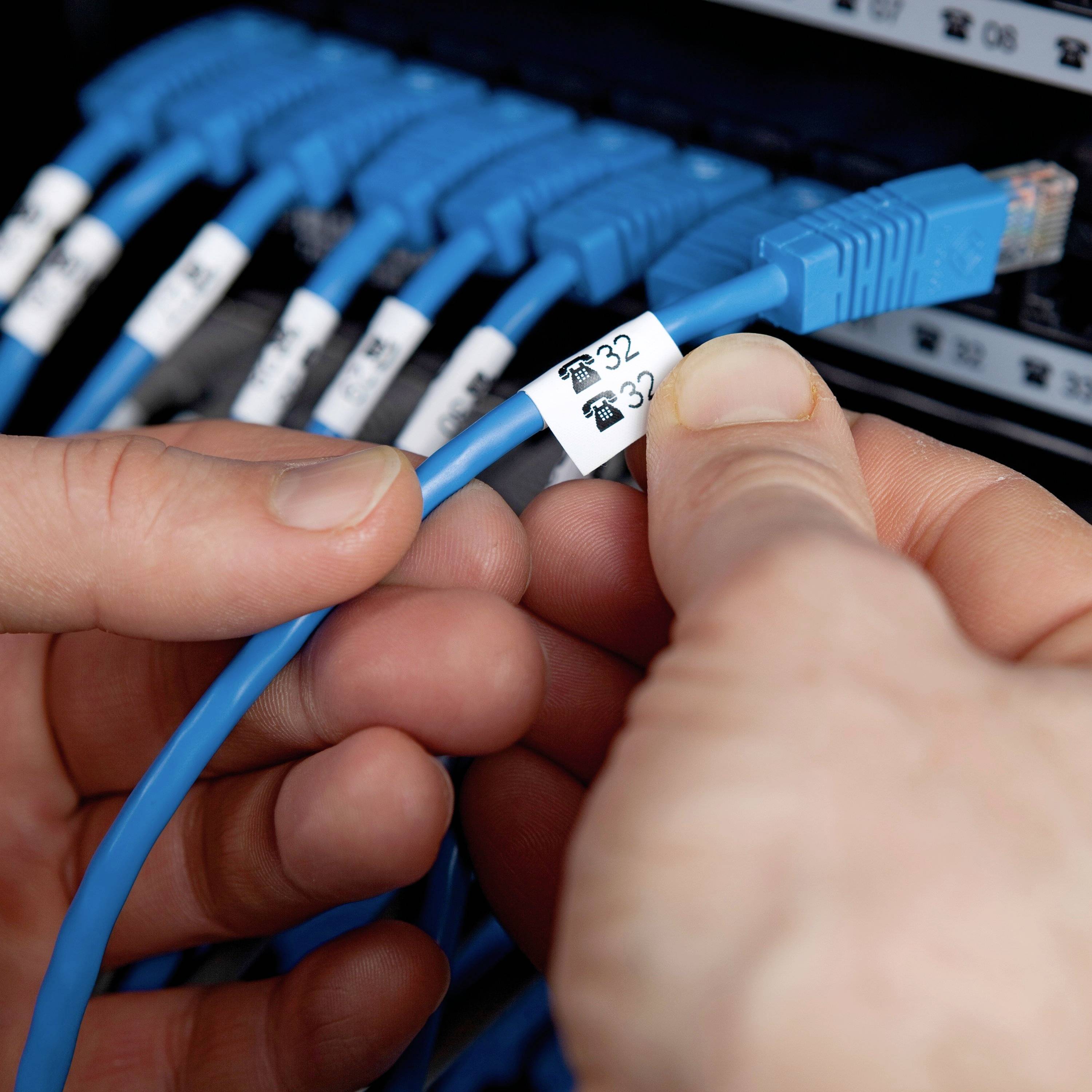Menschliche Hände verbinden ein blaues Netzwerkkabel mit einem Gerät. Ein Etikett am Kabel zeigt Symbole und die Zahl '32'.