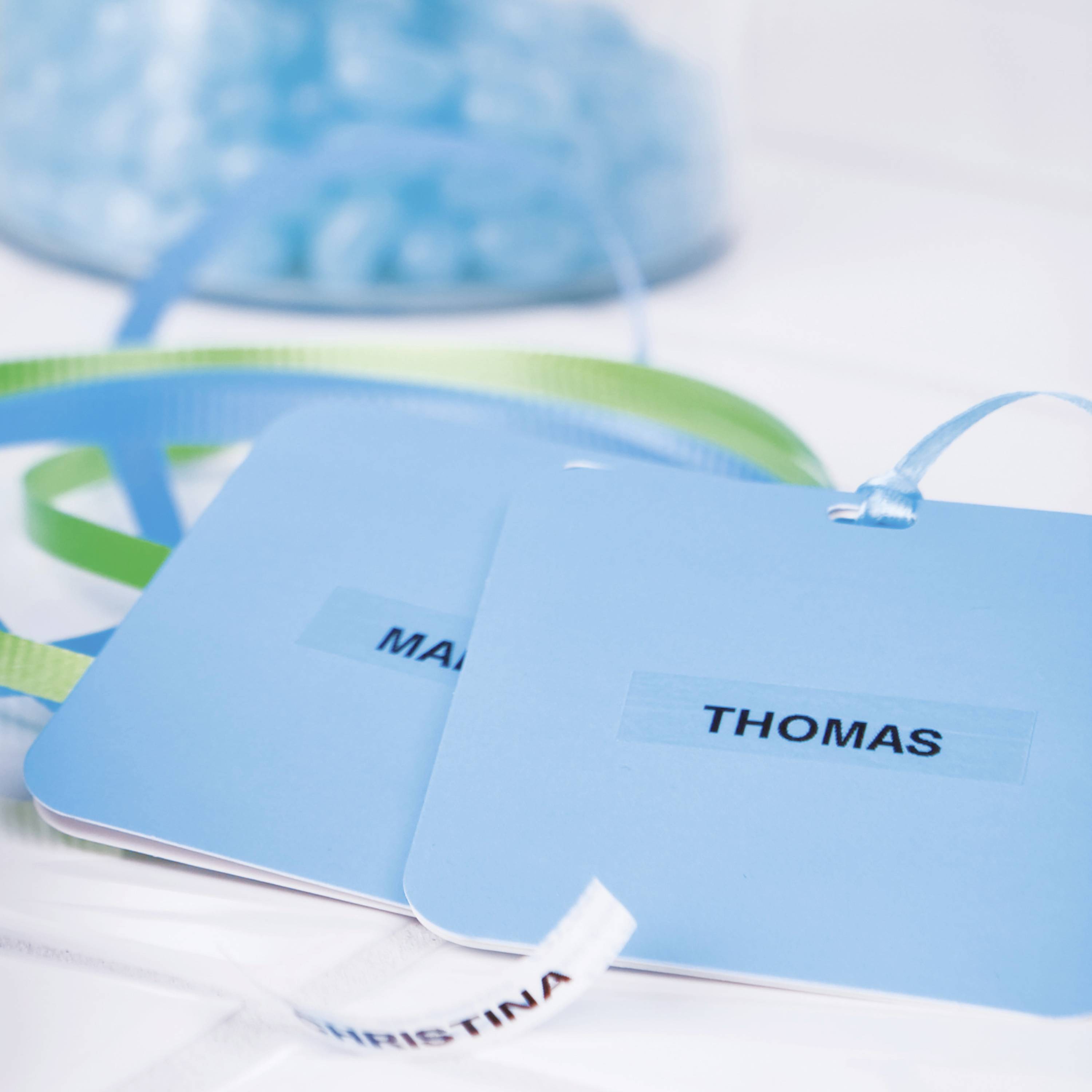 Zwei blaue Namensschilder mit den Namen 'MARIA' und 'THOMAS' liegen auf einem Tisch. Im Hintergrund sind blaue Glassteine zu sehen.