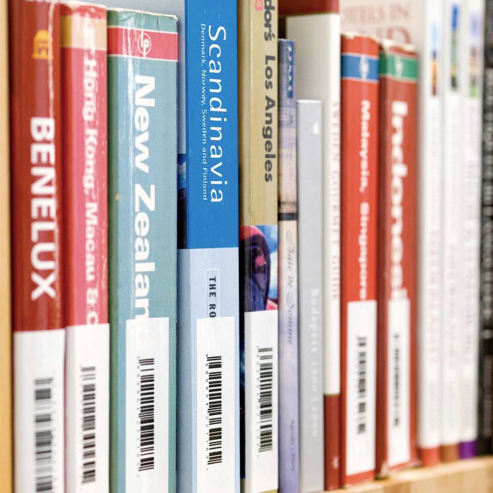 Eine Bibliotheksregalreihe mit Reiseführern zu verschiedenen Ländern und Regionen, darunter Benelux, Neuseeland, und Skandinavien.