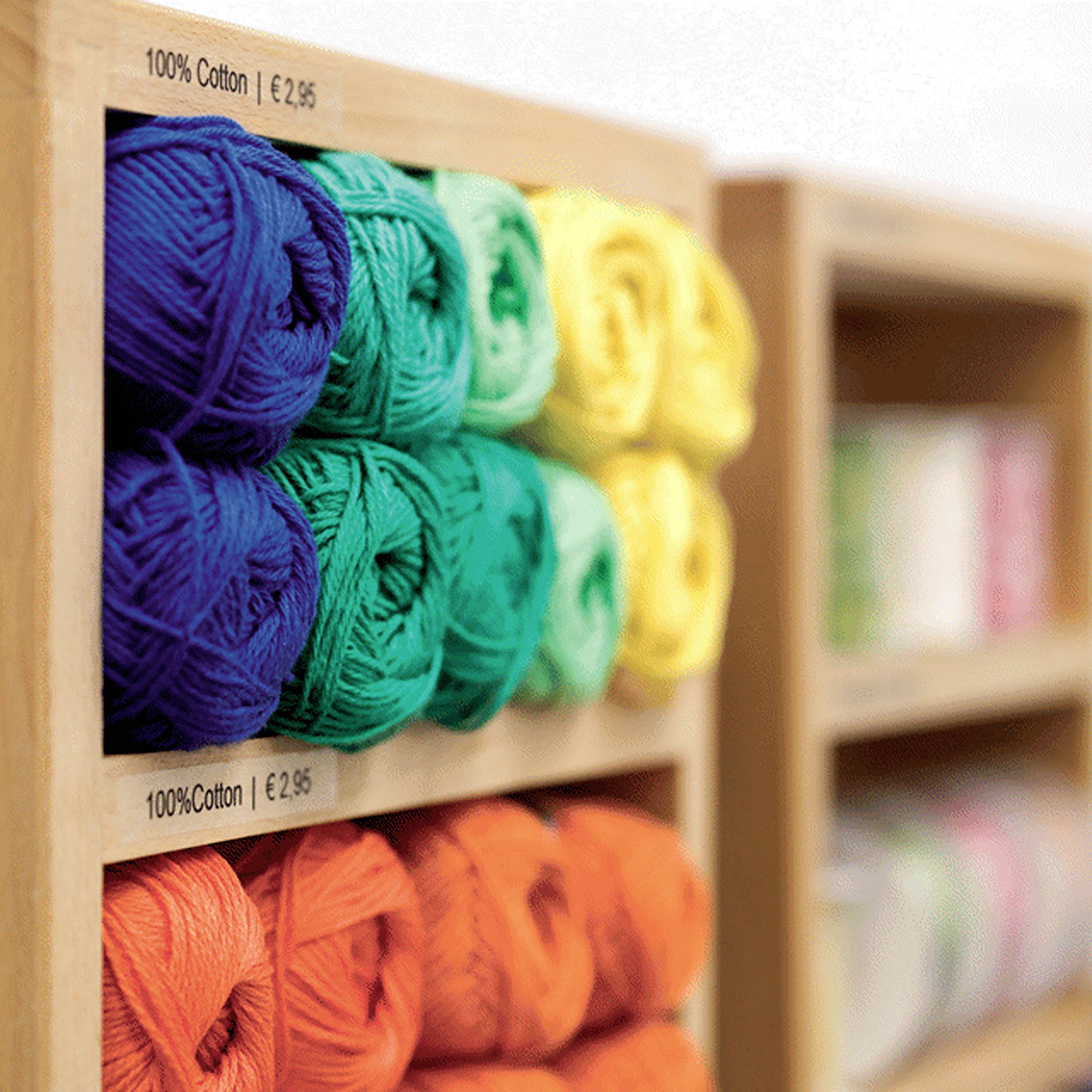Eine Holzregal zeigt bunte Wollknäuel in verschiedenen Farben. Zwei Preisschilder zeigen '100% Cotton' und 'EUR 2.95'.