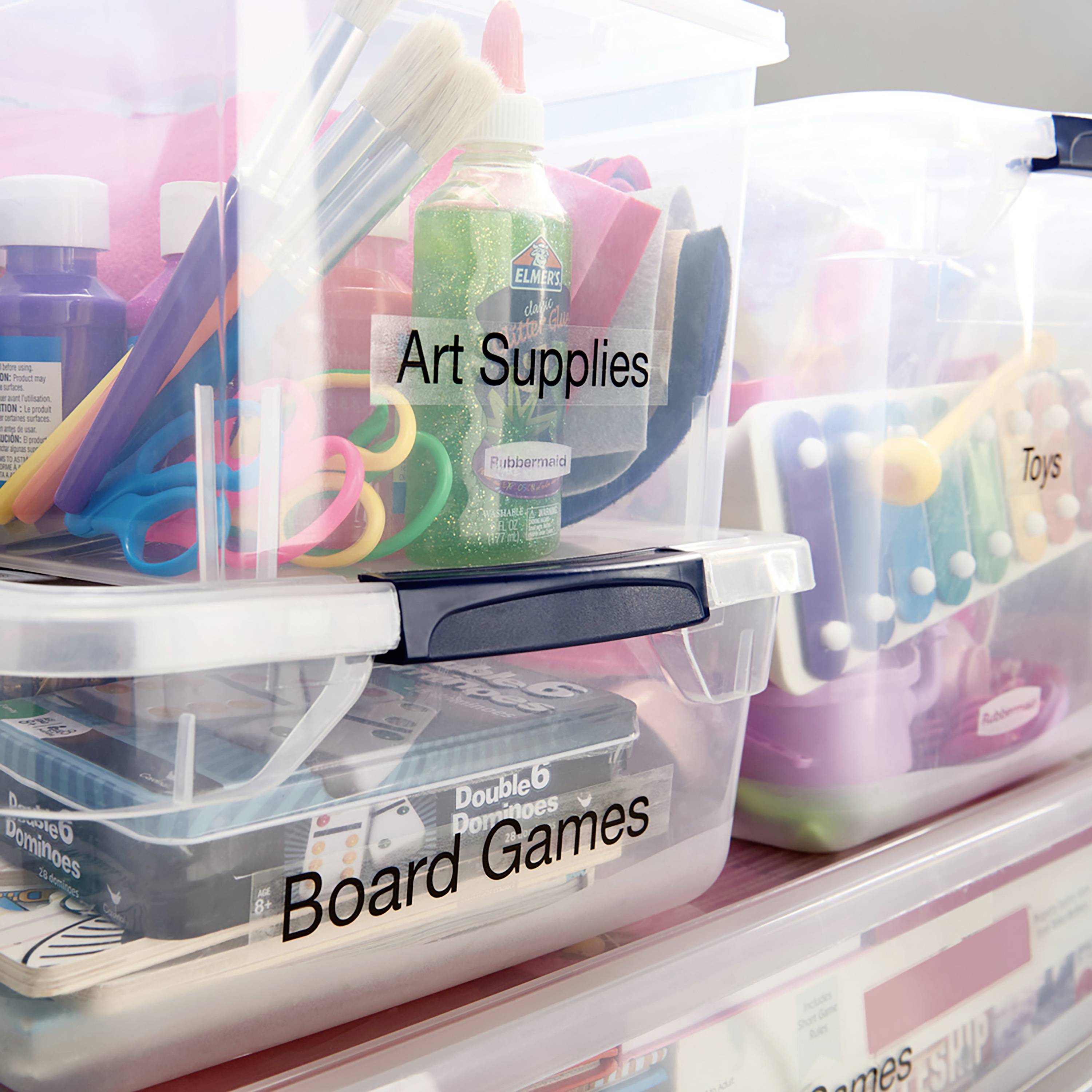Plastikboxen mit Etiketten: ‚Art Supplies‘ mit Pinseln und Farben, ‚Toys‘ mit einem Xylophon, ‚Board Games‘ mit Dominosteinen.