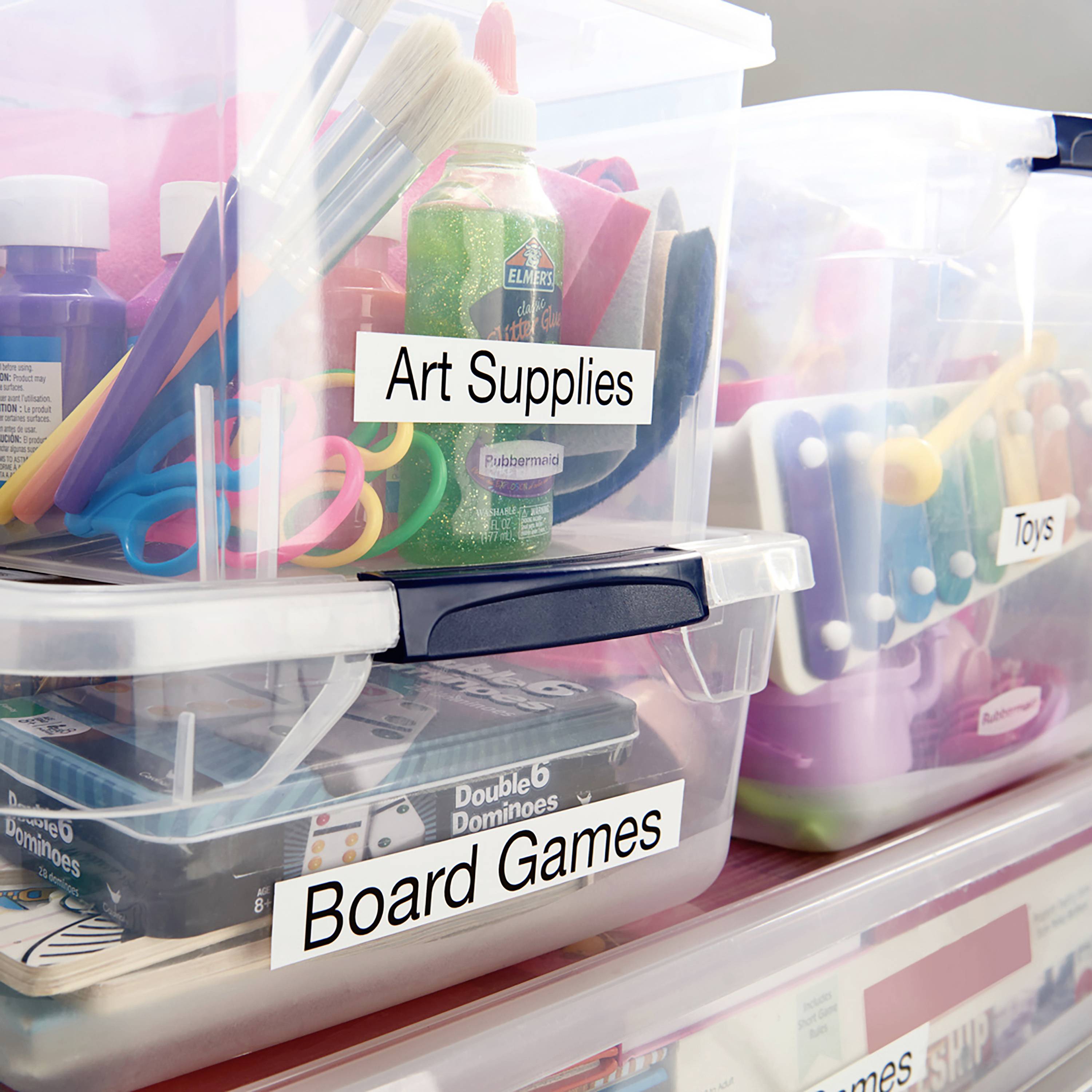 Kunst- und Spielsachen in Plastikboxen. Eine Box mit 'Art Supplies' enthält Pinsel und Farben, die andere mit 'Board Games' enthält Domino.