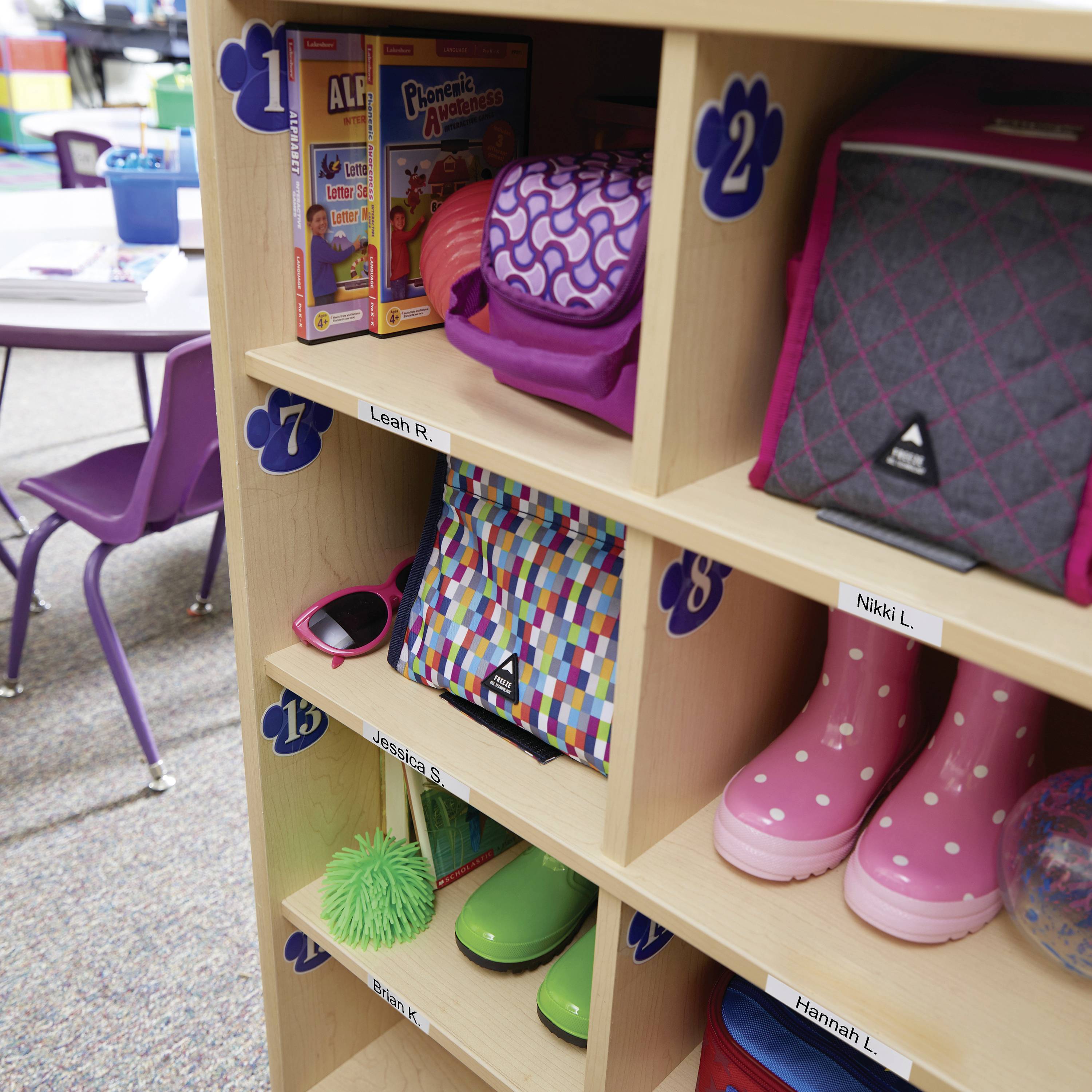 Ein Regal in einem Klassenzimmer mit Schließfächern. Schulsachen wie Bücher, Lunchbox, Rucksack, Sonnenbrille, Gummistiefel und Spielzeug sind in verschiedenen Fächern verstaut.
