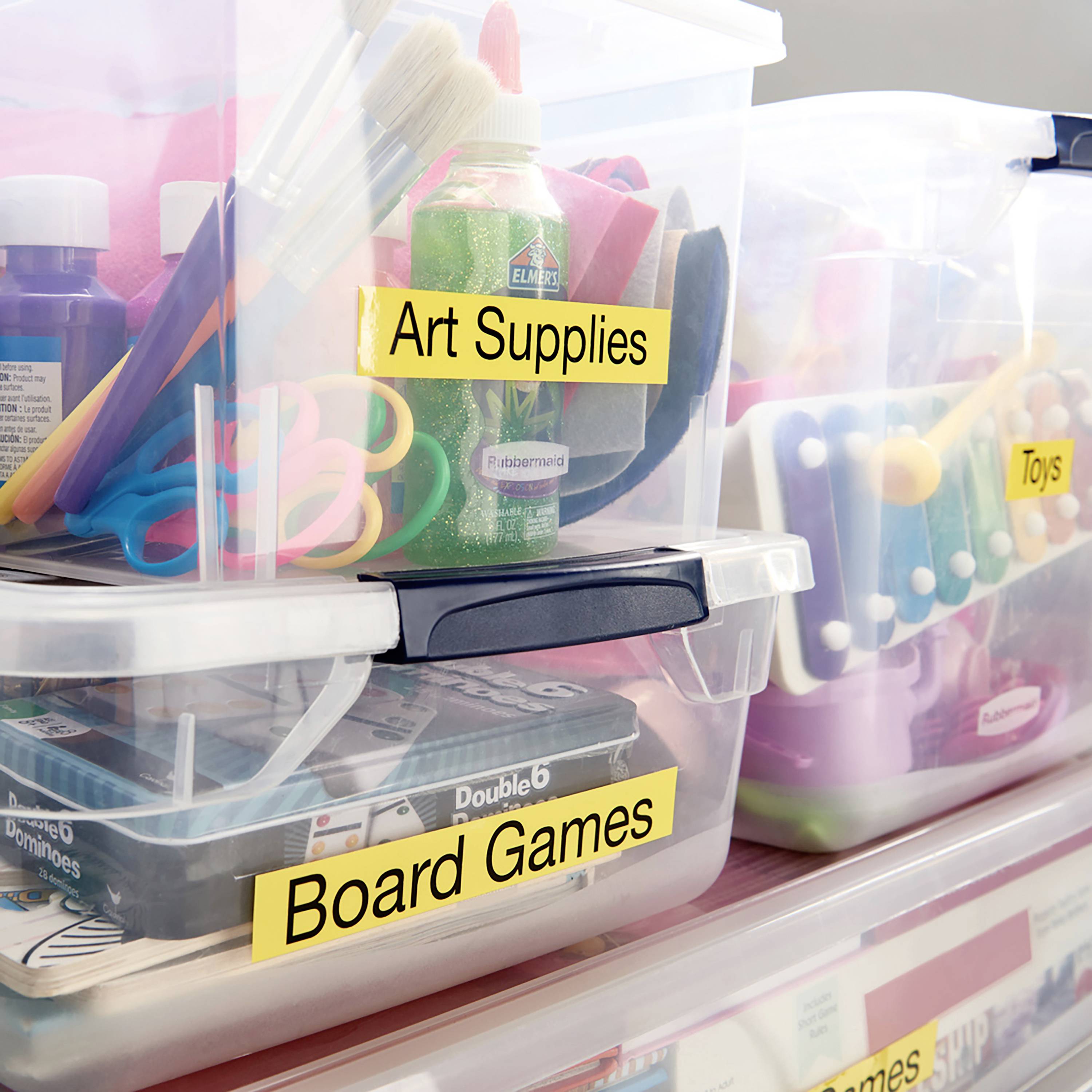 Kunstbedarf, Brettspiele und Spielzeug in transparenten Boxen mit Etiketten, organisiert auf einem Regal.