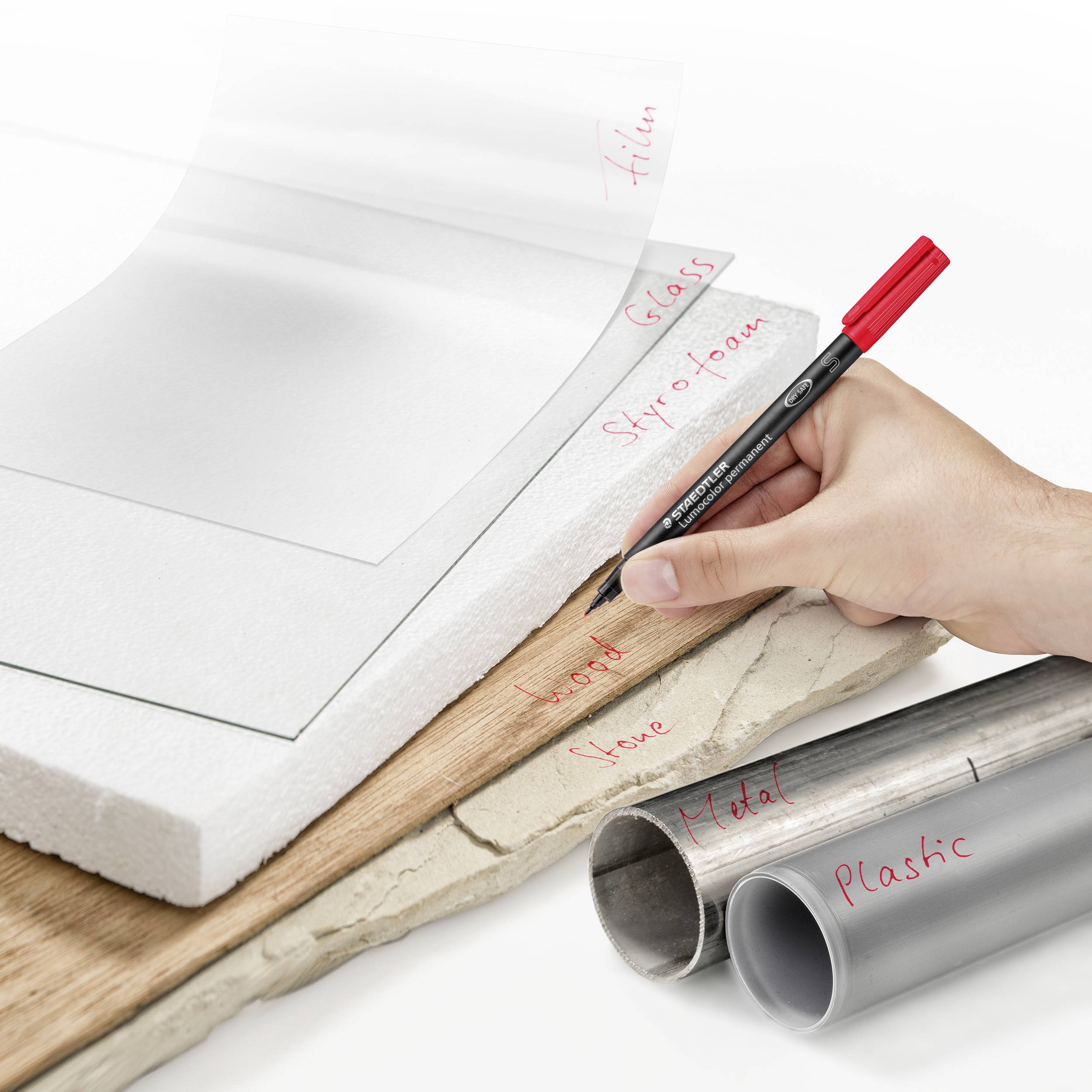 Eine Hand schreibt mit einem roten Stift auf eine Papierschicht, die auf mehreren Materialien wie Holz, Glas, Styropor und Metall liegt.