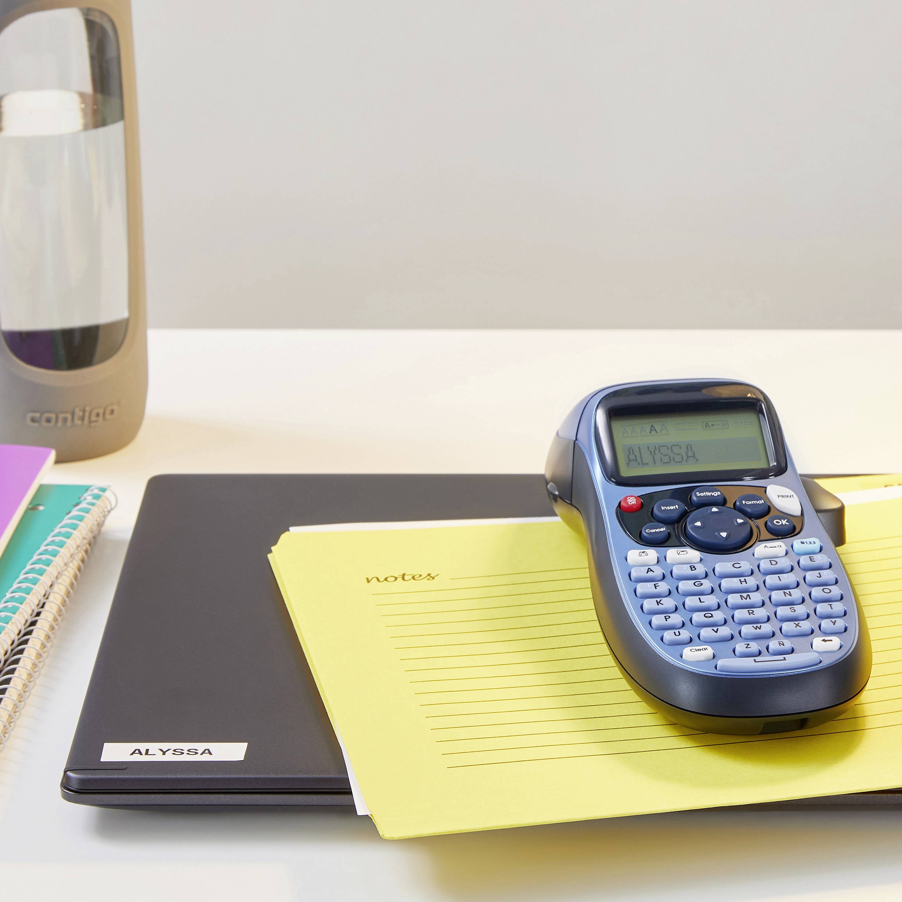 Notizbuch, gelber Block mit 'notes', Etikettendrucker und Thermobecher auf Schreibtisch.