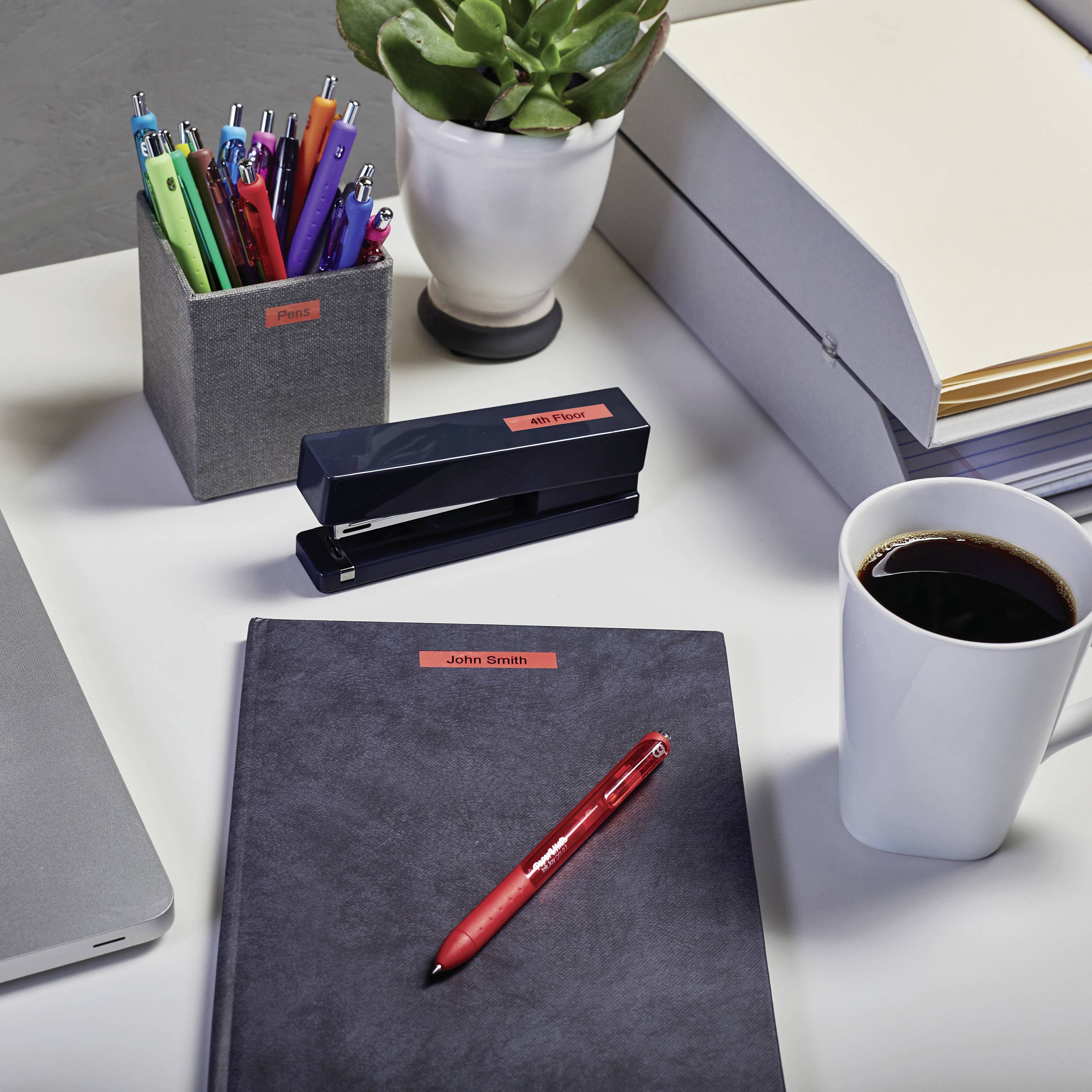 Ein Schreibtisch mit Laptop, Stiften, Pflanze, Taschentücherbox, Kaffeetasse und Notizbuch mit rotem Stift, typisches Büro-Setup.