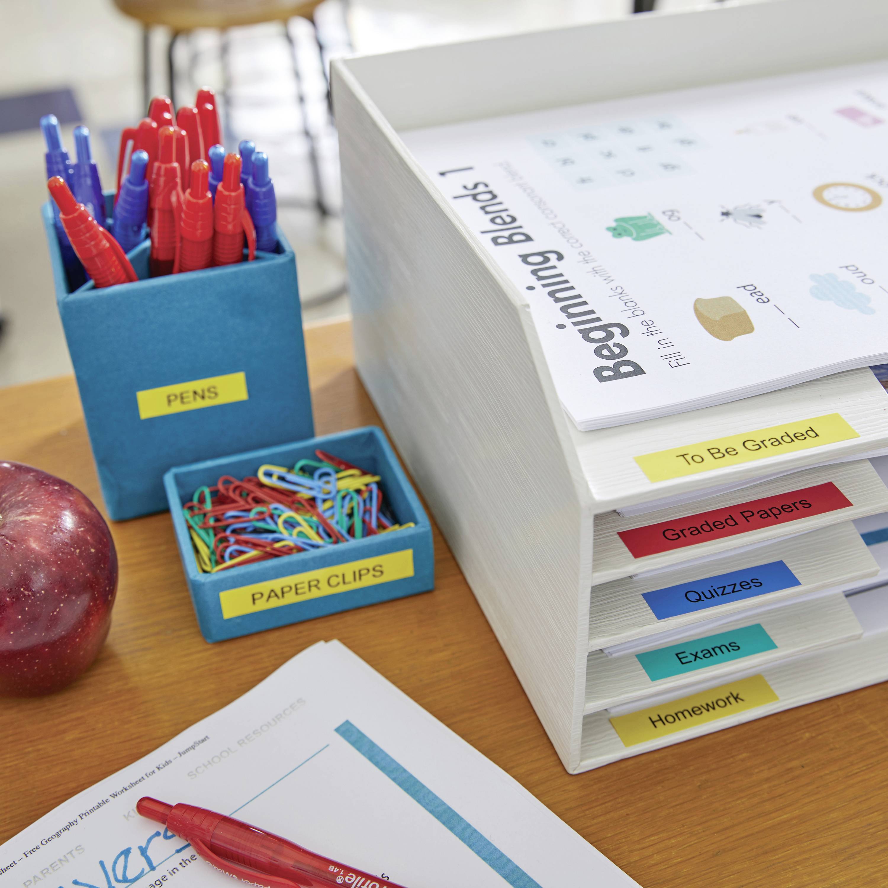 Ein Tisch mit Ordnern aufgestapelt, Etiketten: 'To Be Graded', 'Graded Papers', 'Exams', 'Quizzes', 'Homework'. Stifte und Büroklammern daneben.