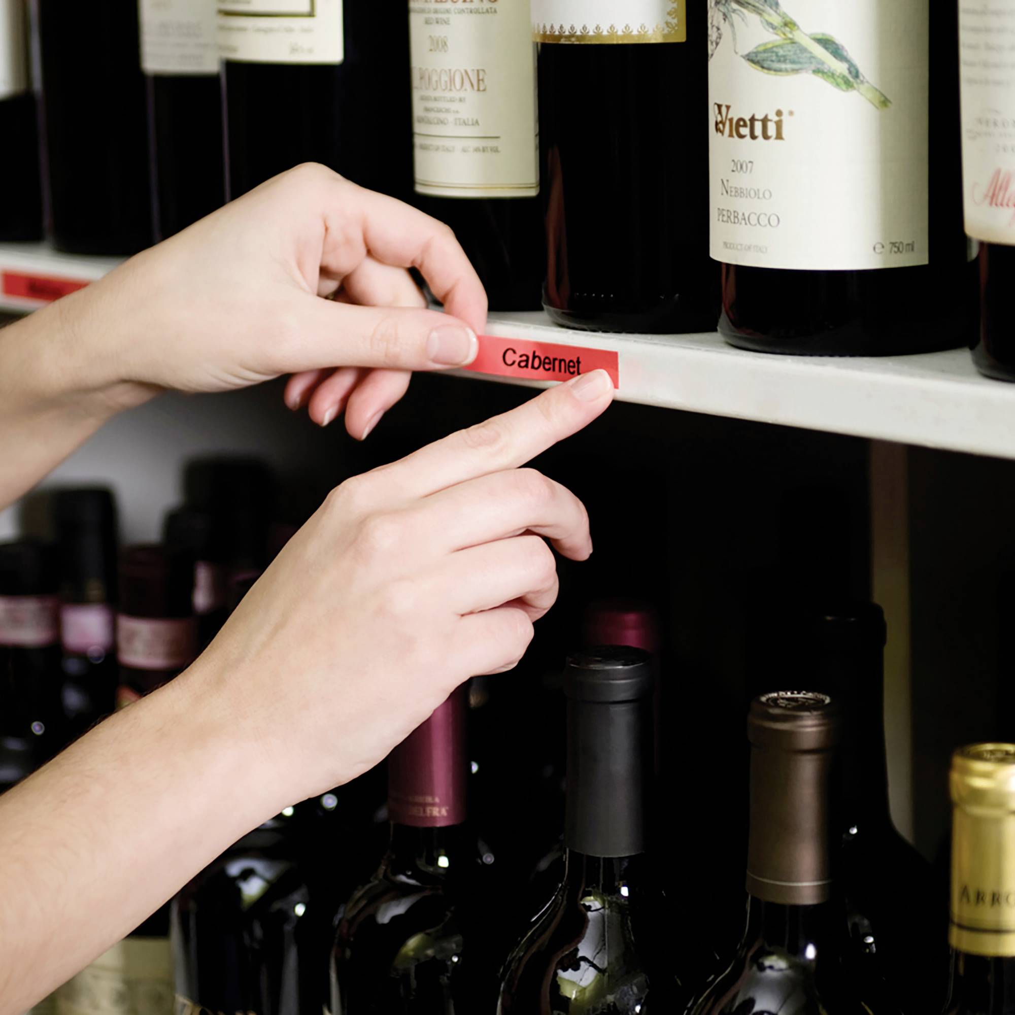 Eine Hand platziert ein rotes Etikett mit der Aufschrift 'Cabernet' auf einem Weinregal, um Flaschen zu kennzeichnen. Im Hintergrund sind Weinflaschen sichtbar.