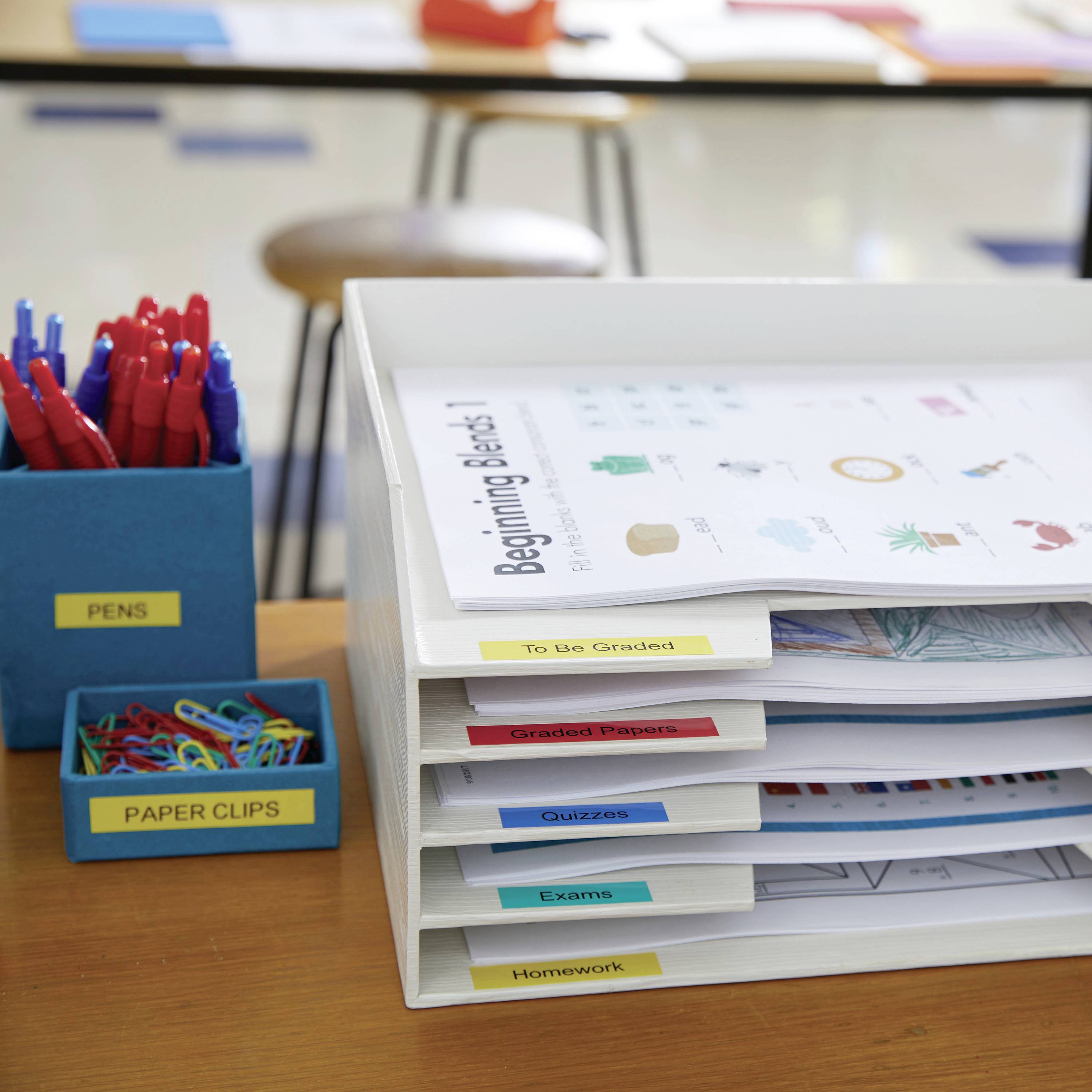 Ein Schreibtisch mit einem Ablagesystem für Papiere, markiert mit Labels wie 'To Be Graded', neben einem Behälter mit Stiften und Büroklammern.