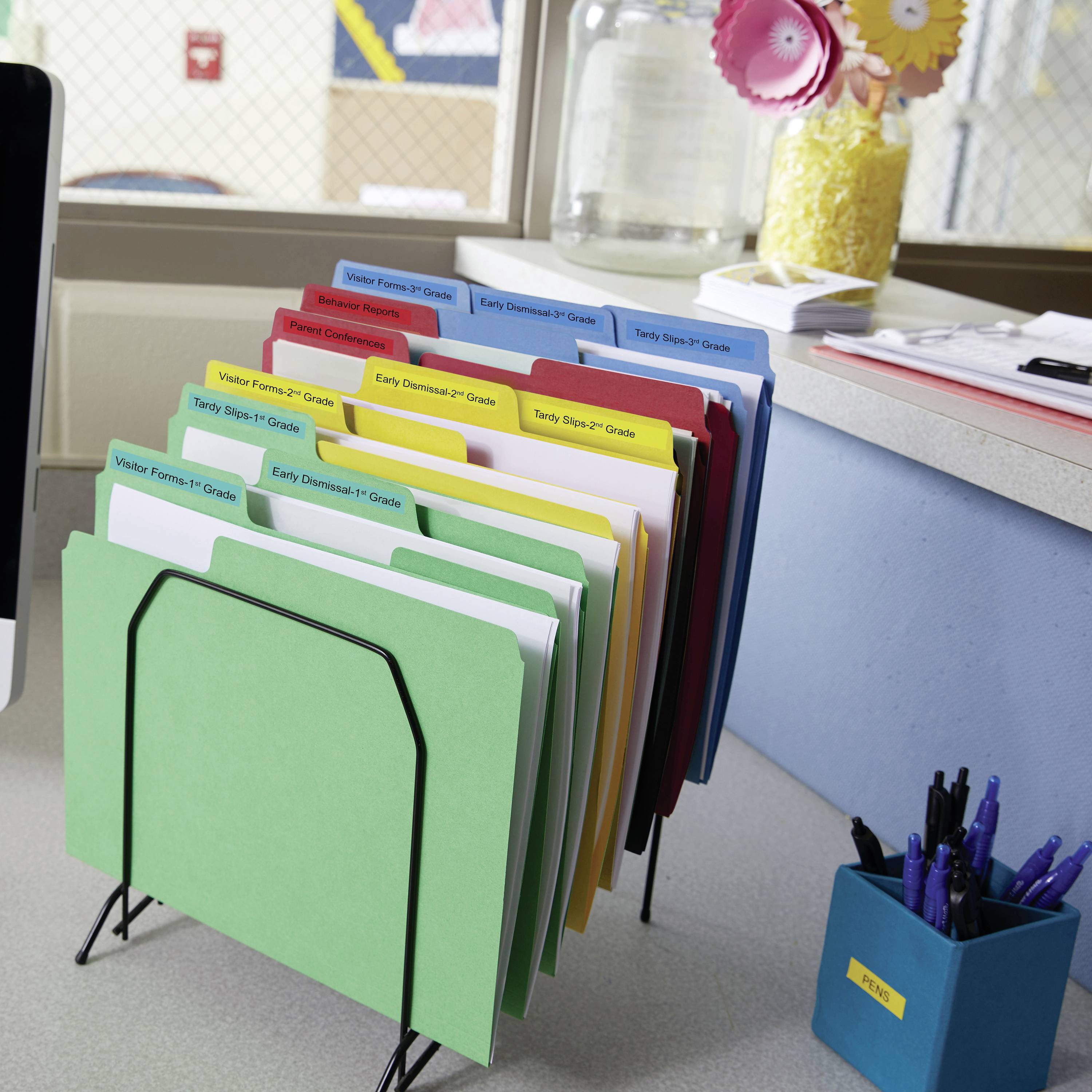 Ein Schreibtisch mit einem Aktenhalter, der bunte Ordner mit Beschriftungen für verschiedene Schulstufen enthält. Im Hintergrund ein Glas mit dekorativen Blumen.