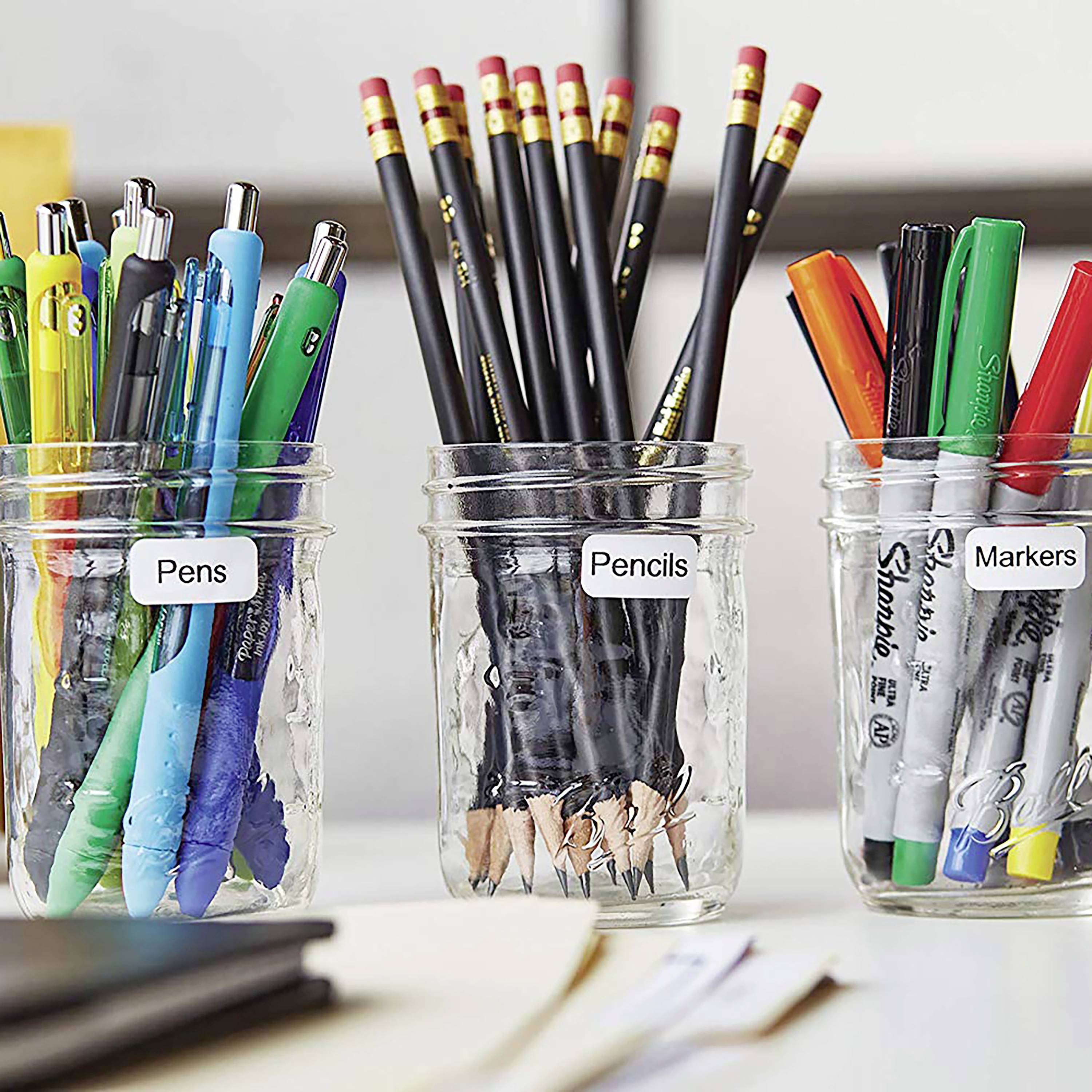 Drei Glasgefäße mit Beschriftungen 'Pens', 'Pencils', 'Markers' stehen auf einem Tisch, gefüllt mit unterschiedlichen Schreibutensilien.