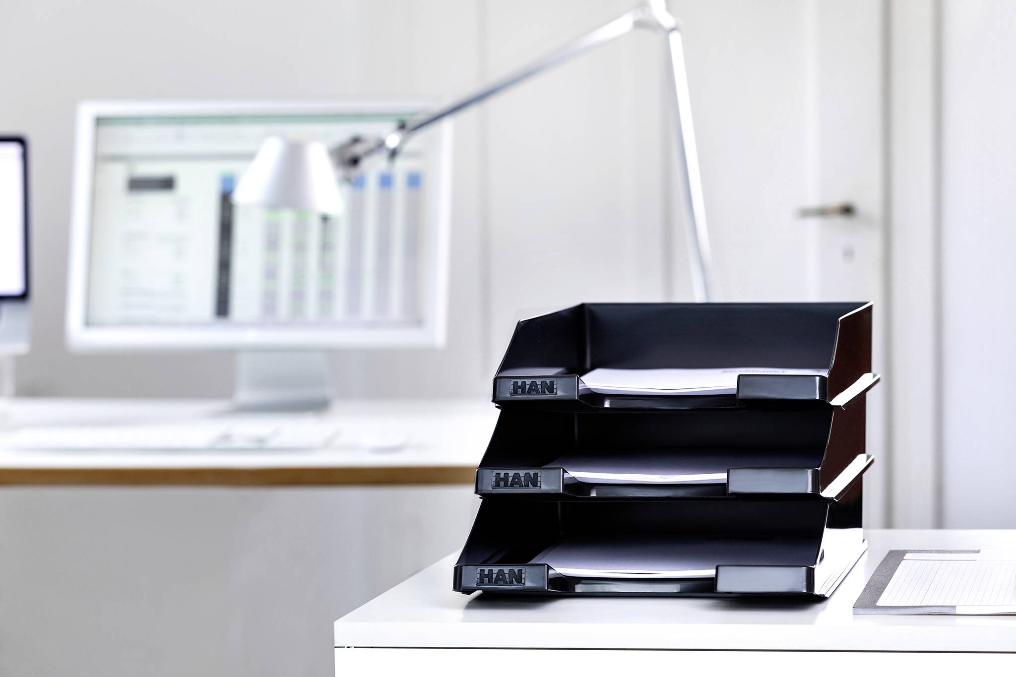 Ein moderner Arbeitsplatz mit drei schwarzen Ablagefächern auf einem Schreibtisch, im Hintergrund ein Monitor und eine Lampe.