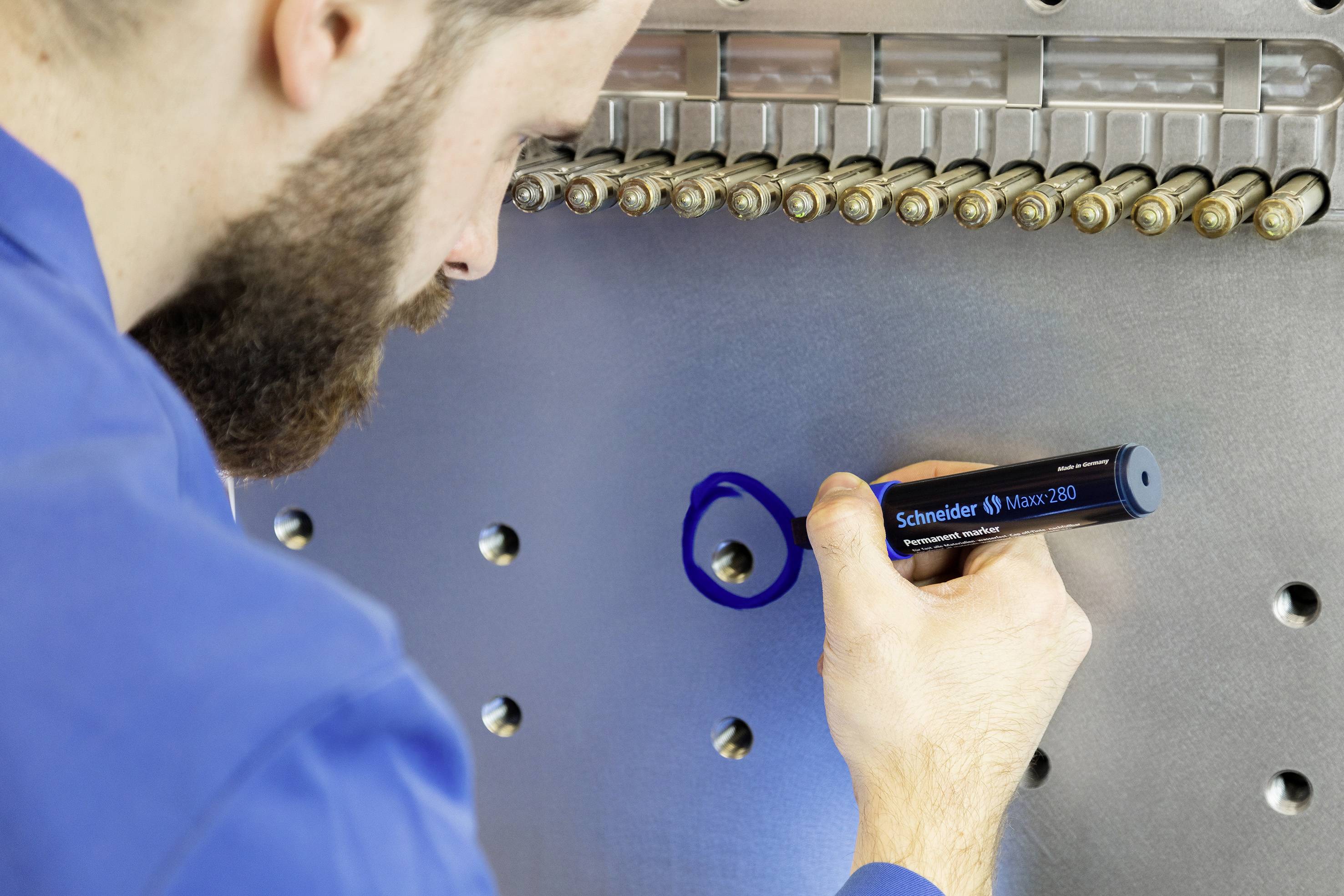 Ein Mann in blauer Arbeitskleidung markiert mit einem blauen Stift eine Metalloberfläche. Mehrere Nieten oder Bolzen sind sichtbar.