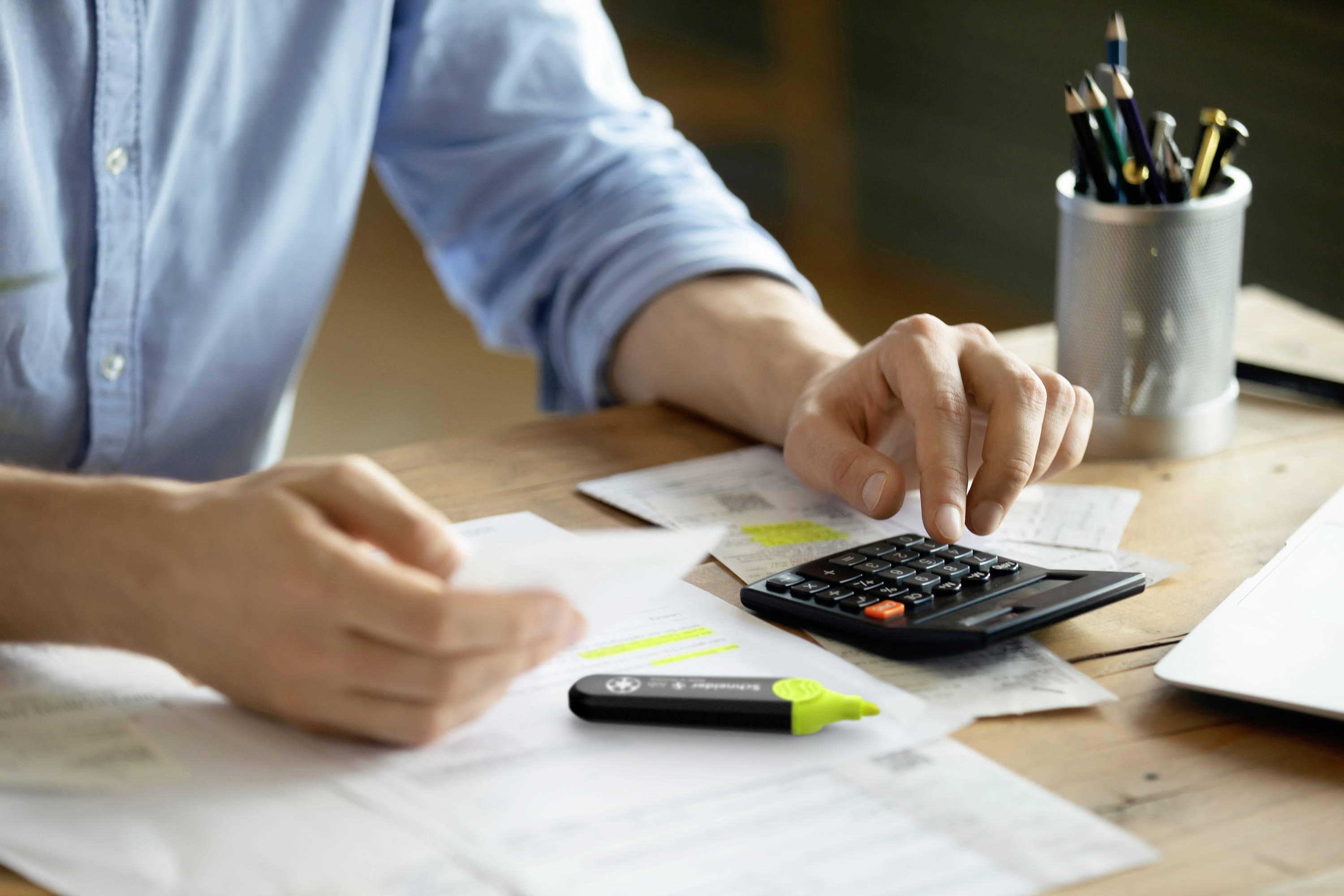 Eine Person benutzt einen Taschenrechner auf einem Tisch voller Papierkram und Unterlagen, vermutlich bei der Arbeit an Finanzen oder Budgetierung.
