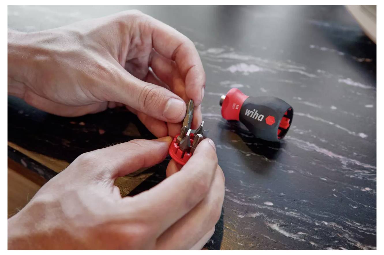 Hände stellen einen roten Werkzeugbithalter mit mehreren austauschbaren Bits ein; ein kleiner schwarz-roter Schraubendrehergriff liegt in der Nähe auf einer dunklen Oberfläche.