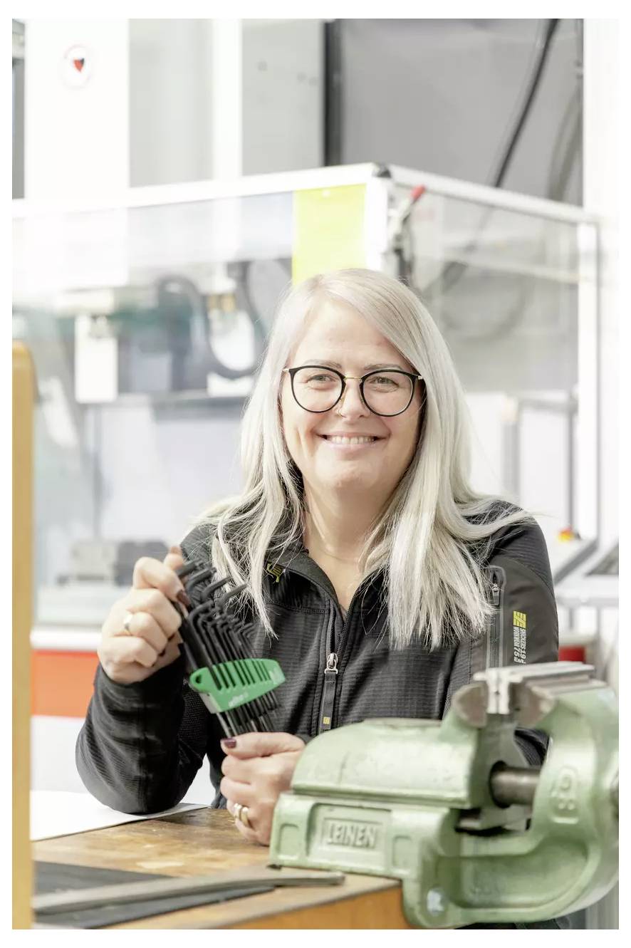 Eine Person mit langen Haaren und Brille lächelt, während sie ein Werkzeug hält. Sie befinden sich in einer Werkstattumgebung, wobei Ausrüstung im Hintergrund sichtbar ist.