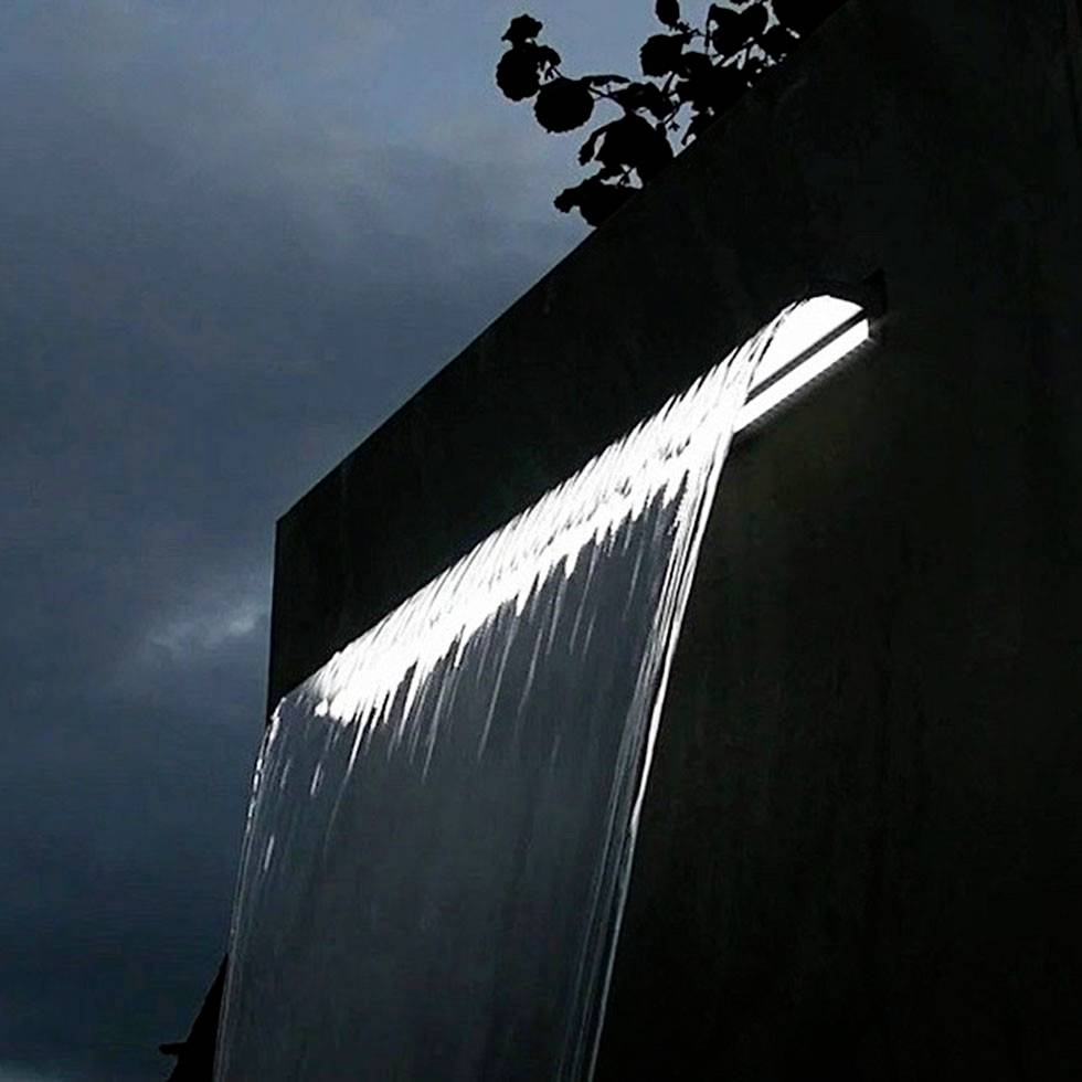 Ein Wasserfall fließt seitlich von einem rechteckigen Betonbau, von düsterem Himmel und Laubwerk eingerahmt, im Licht schimmernd.