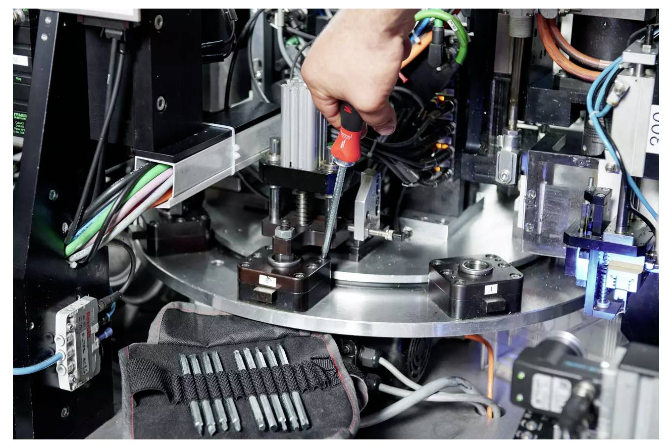 Eine Person verwendet einen Schraubendreher, um Maschinen einzustellen, umgeben von verschiedenen mechanischen Komponenten und Kabeln in einer industriellen Umgebung.