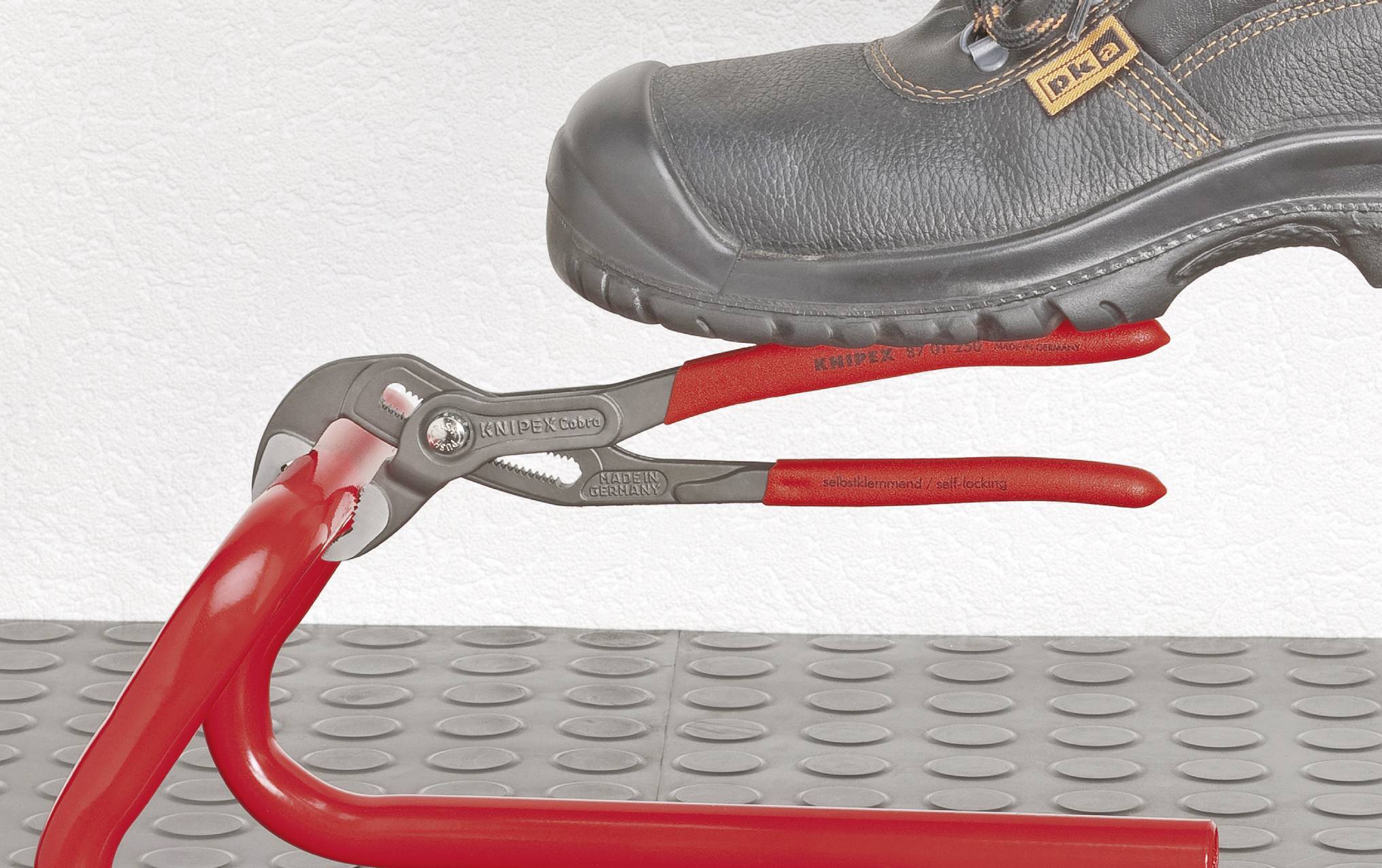 Ein Schuh steht auf einer roten Zange, die ein rotes Rohr hält. Die Zange ist auf einer grauen, strukturierten Fläche platziert.