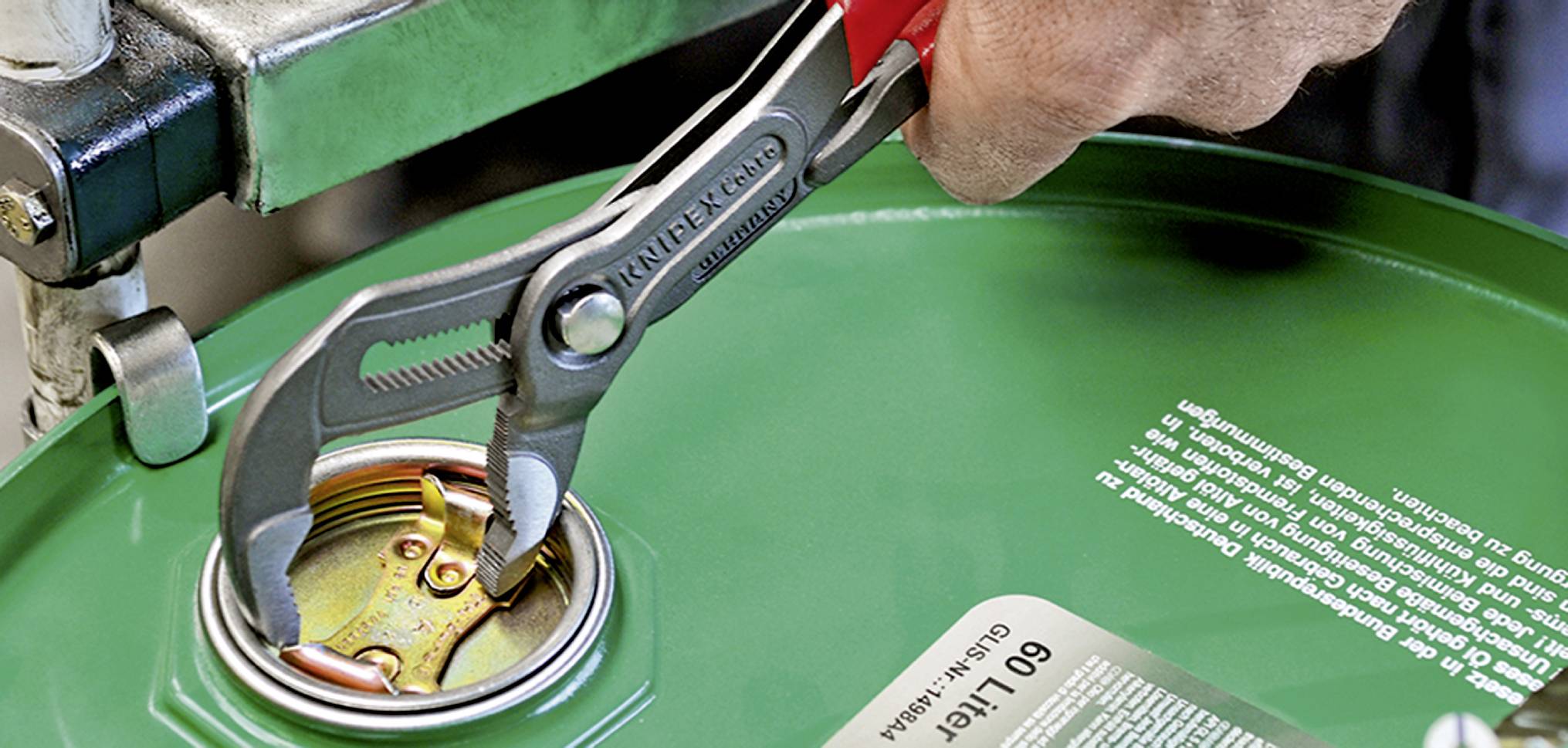 Eine Hand benutzt eine Rohrzange, um den Deckel eines grünen Fasses zu öffnen. Der Deckel ist aus Metall und die Hand wirkt kräftig.