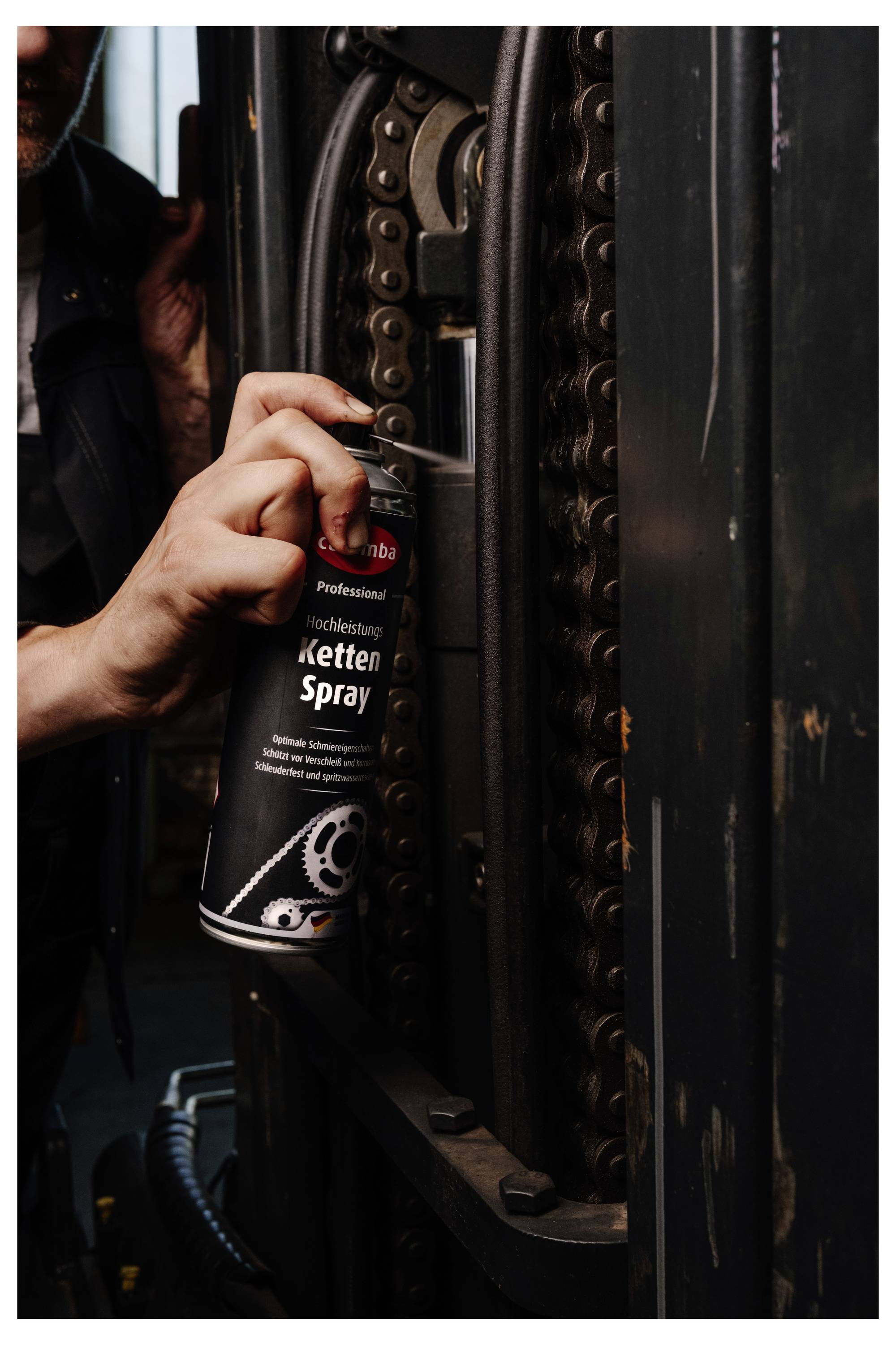 Hand sprays lubricant onto a motorcycle chain with a can labeled 'Ketten Spray'.