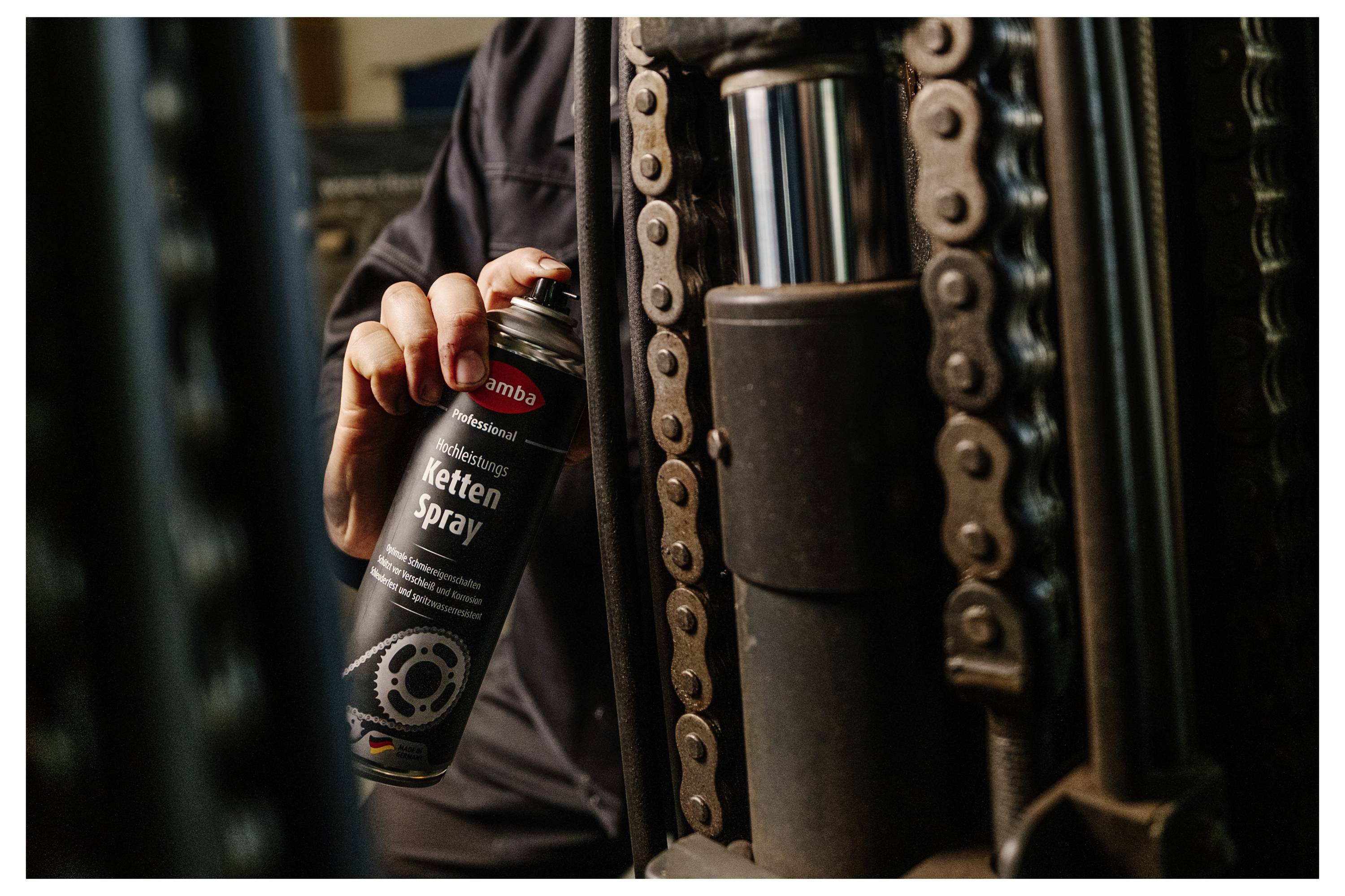Person spraying chain lubricant labeled 'Ketten Spray' onto an industrial machine chain close-up.