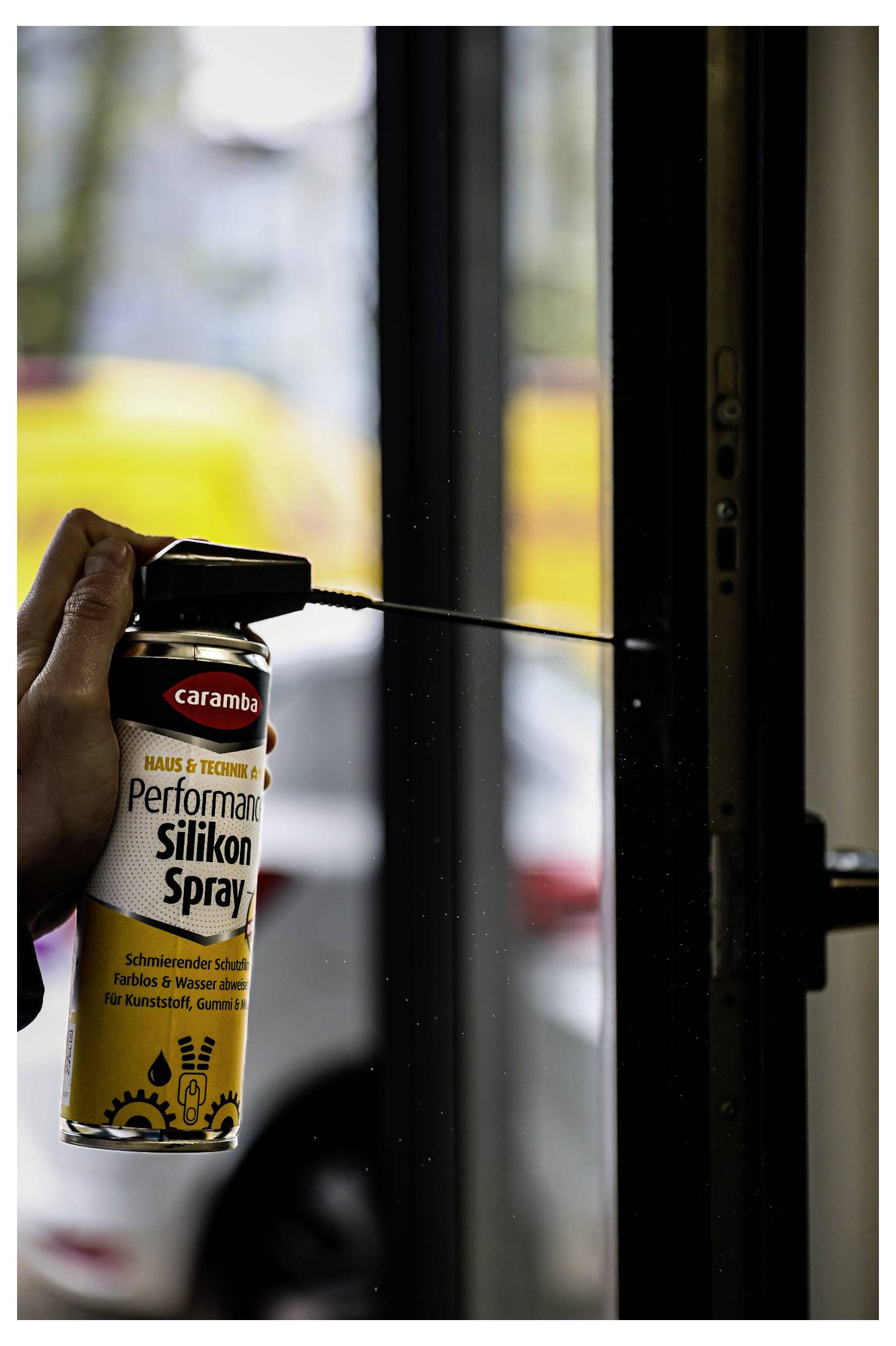 Eine Hand sprüht Silikonschmiermittel aus einer Caramba-Sprühdose auf eine Fenster- oder Türschiene, wobei die schwarze Düse auf den Rahmen gerichtet ist.