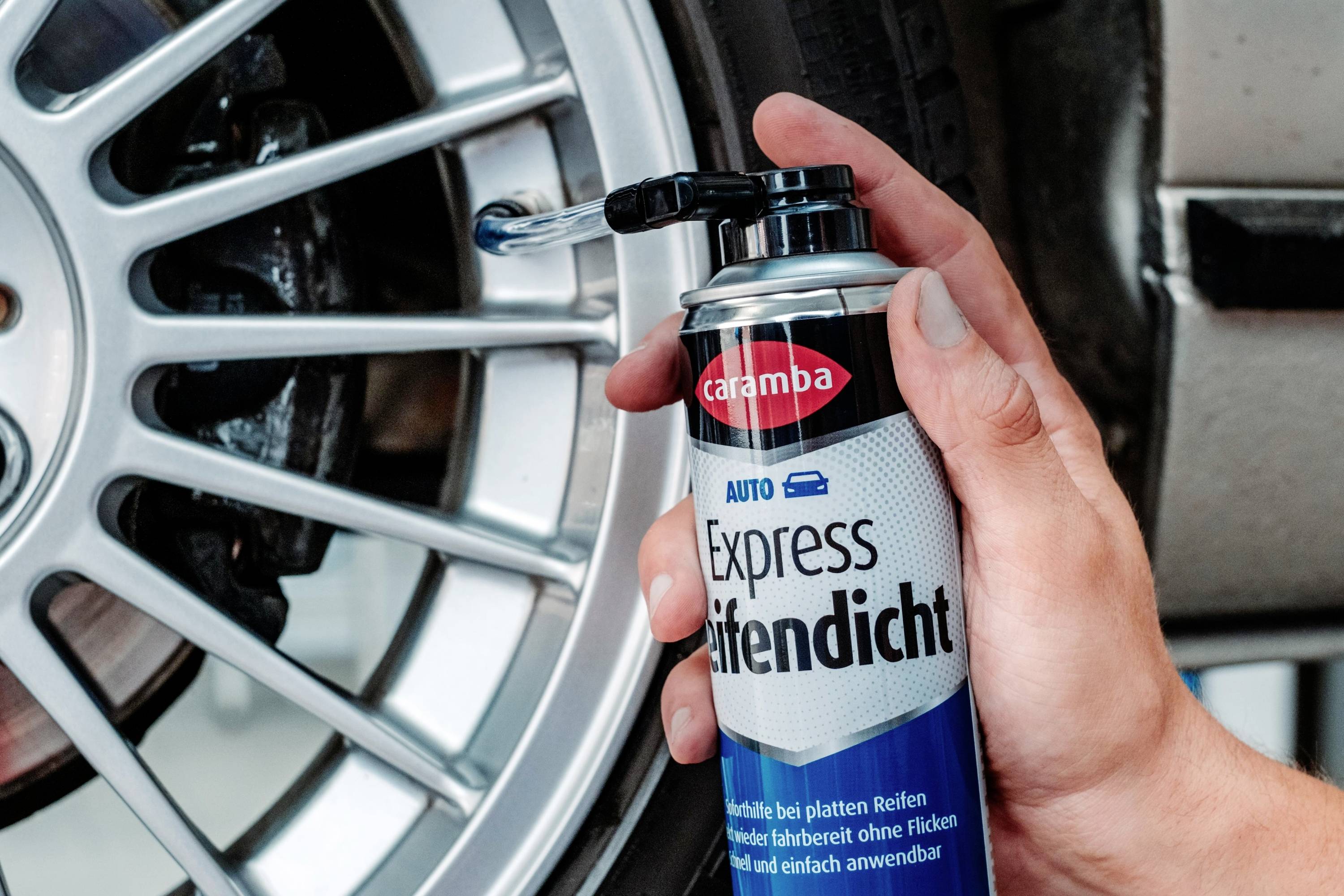 Eine Hand hält eine Sprühdose mit der Aufschrift Express Reifendicht in der Nähe eines Autofelgenrands, was auf eine Verwendung zur Reifenwartung oder -reparatur hindeutet.