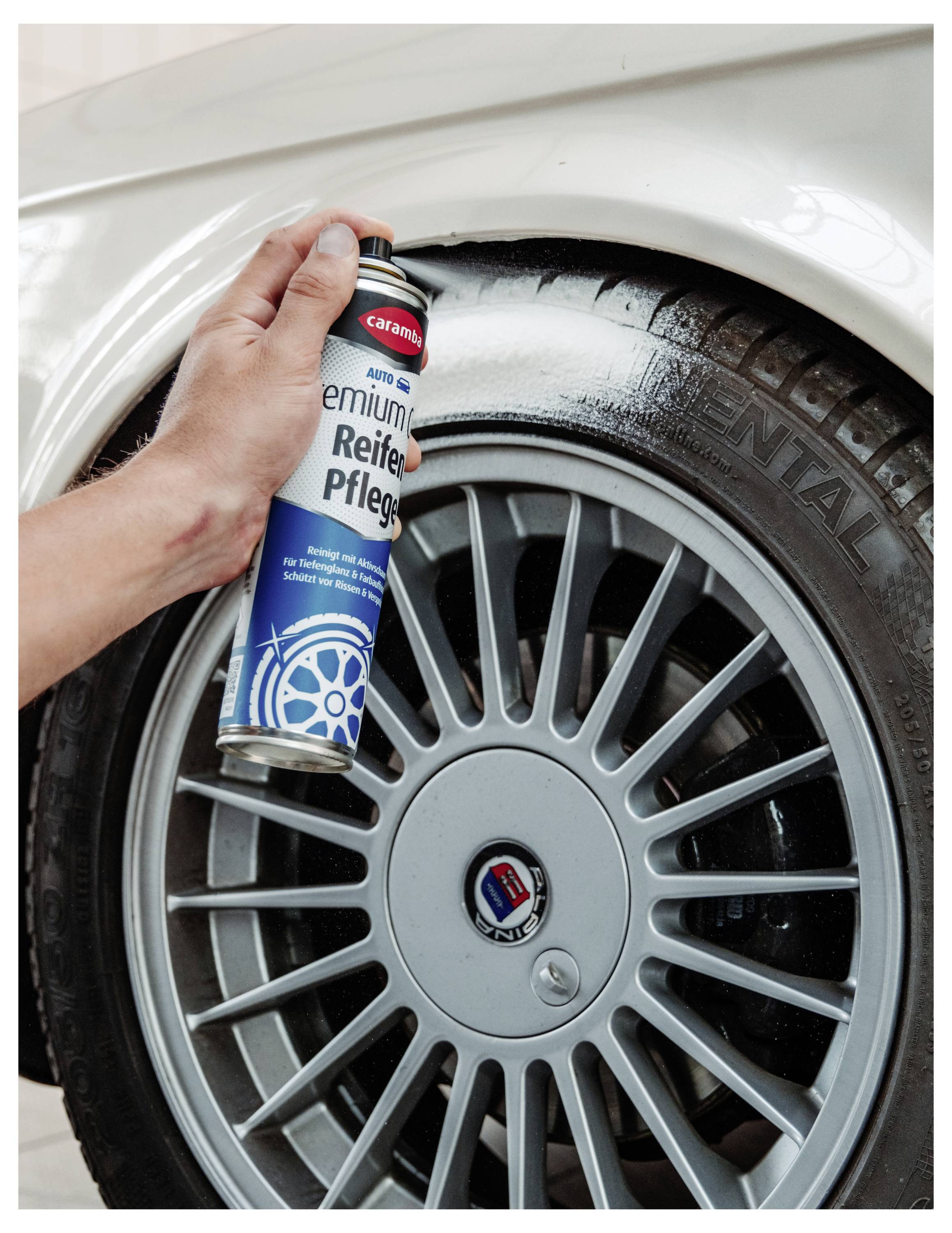 Mit einer Spraydose von Caramba 'Premium Reifen Pflege' wird von Hand Reifenpflege auf ein Autorad und einen Reifen gesprüht.