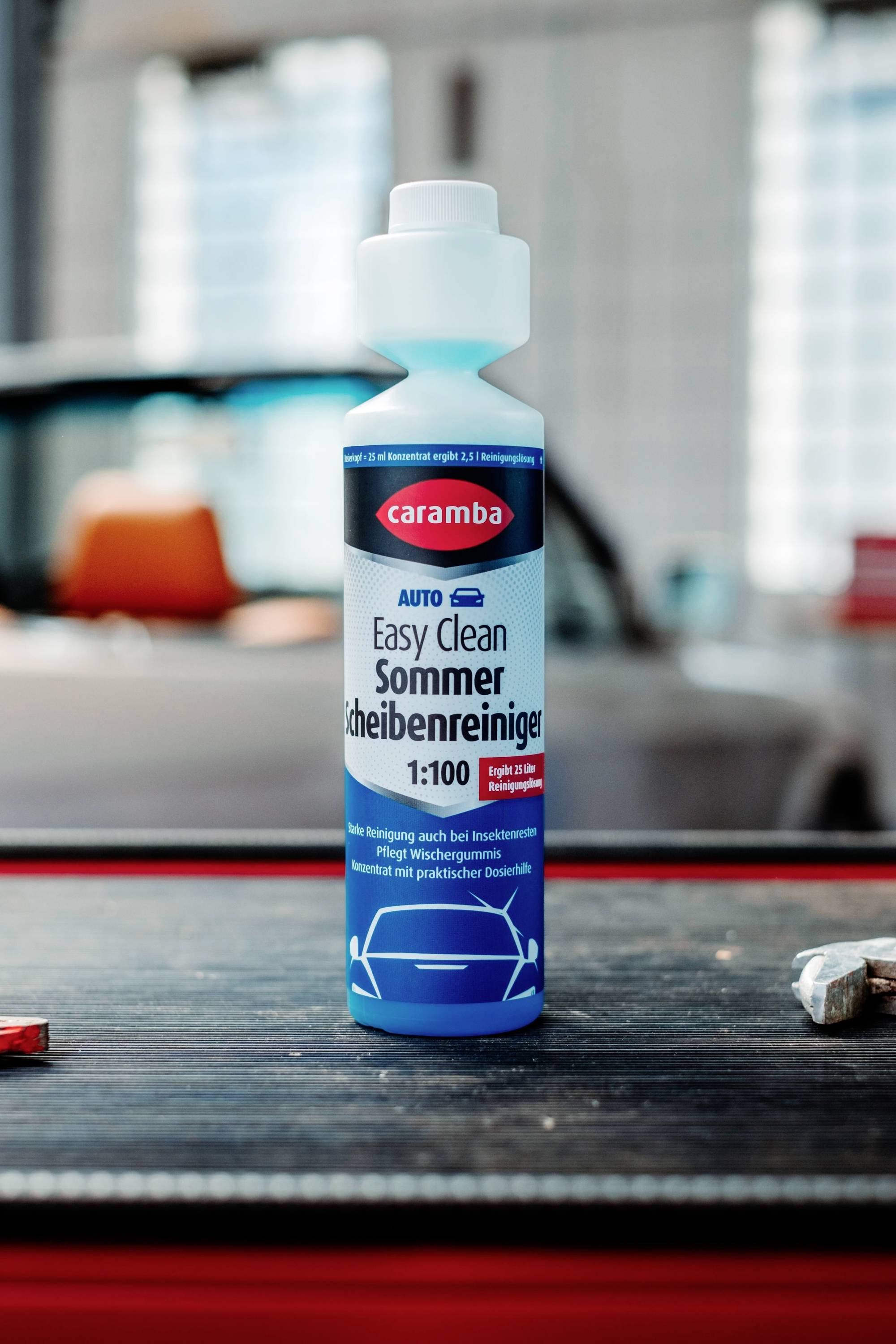 Eine blaue Flasche Auto-Scheibenreiniger mit der Aufschrift 'Easy Clean Summerscheibenreiniger' steht auf einem Tisch, während ein Auto im Hintergrund unscharf zu sehen ist.