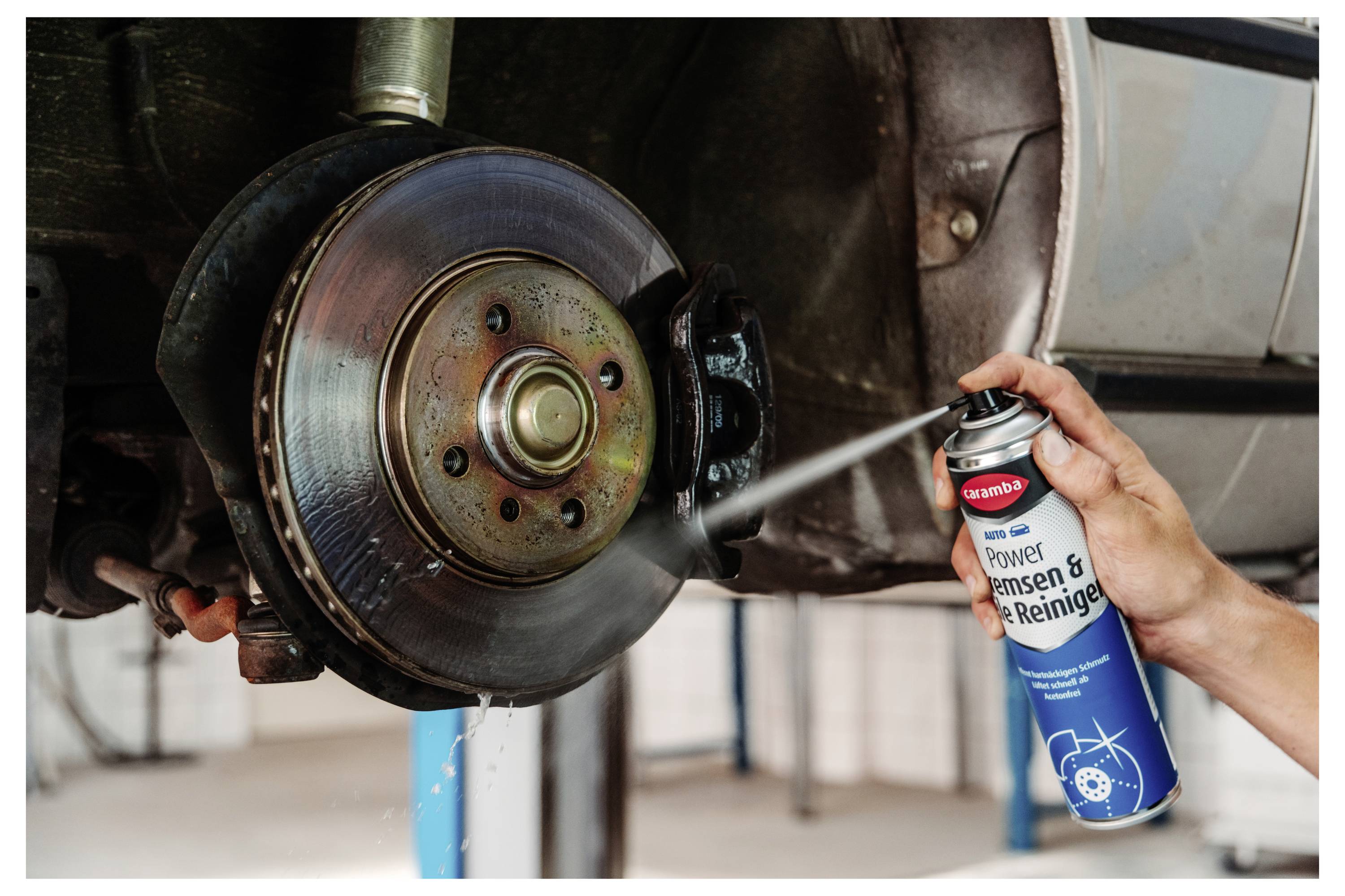 Mechaniker sprüht Bremsreiniger auf einen freiliegenden Bremsrotor und Bremssattel eines Autos während der Wartung.