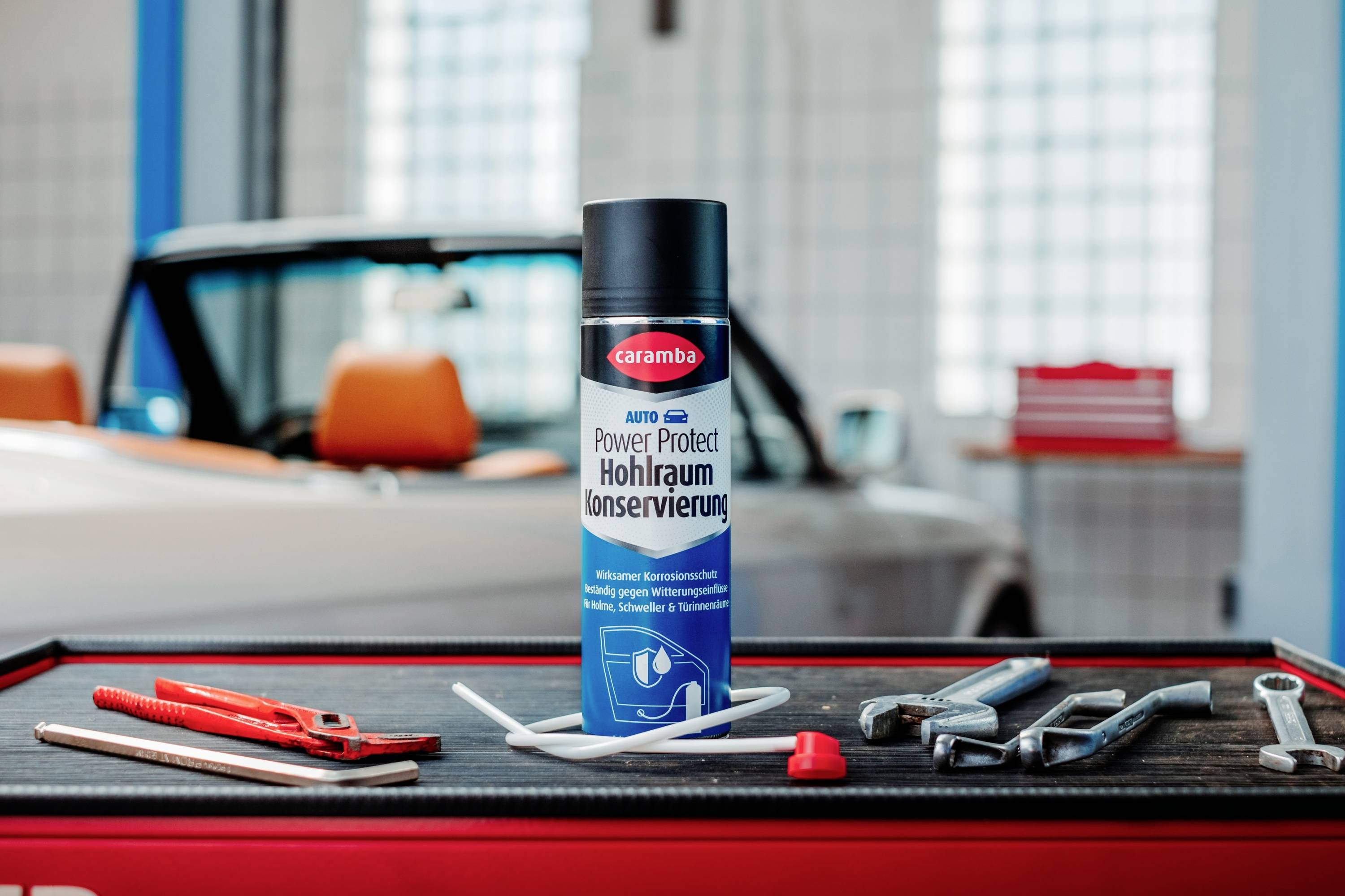 Eine Dose Caramba Auto 'Power Schutz Hohlraumversiegelung' auf einem Tisch mit Autowerkzeugen, vor einem klassischen Cabrio.