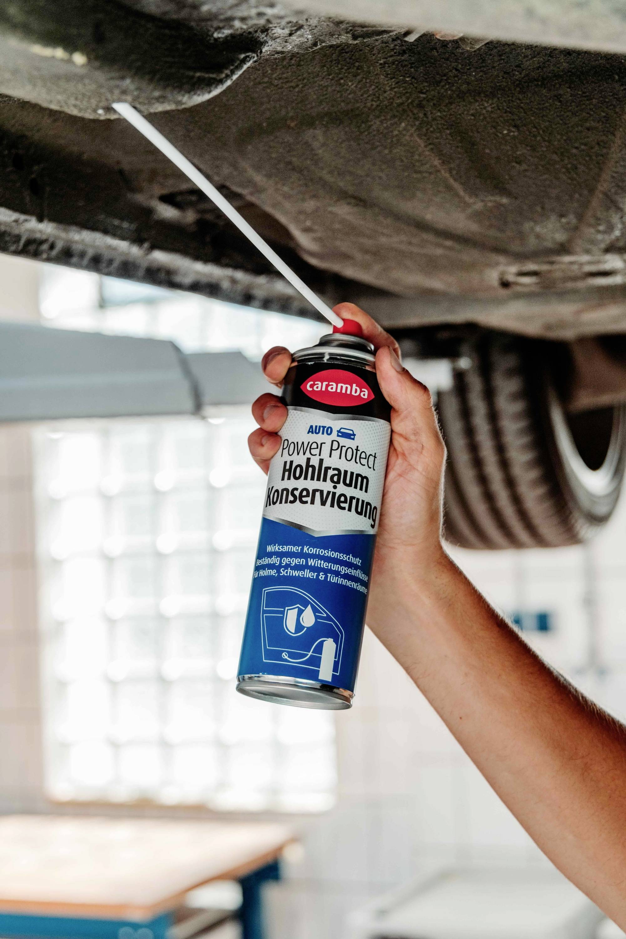 Eine Person trägt Fahrzeugschutzspray an der Unterseite eines Fahrzeugs mit einer Sprühdose mit der Aufschrift 'Power Protect Hohlraum Konservierung' auf.