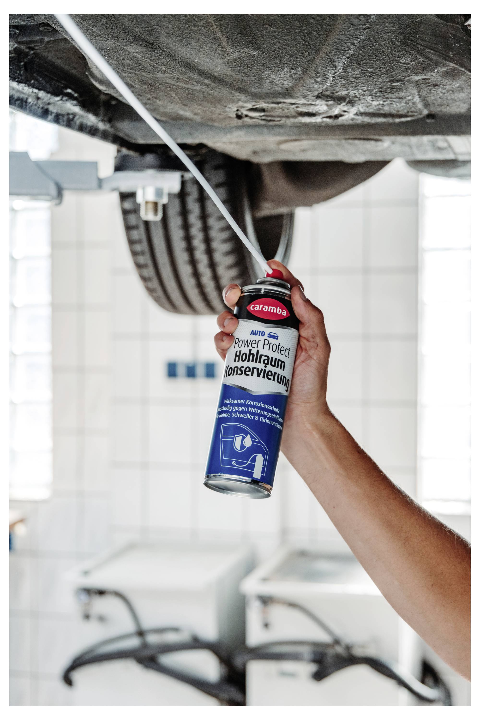 Hand spraying von Schmiermittel aus einer Dose auf die Unterseite eines Autos, während das Fahrzeug in einer Garage oder Werkstatt angehoben ist.