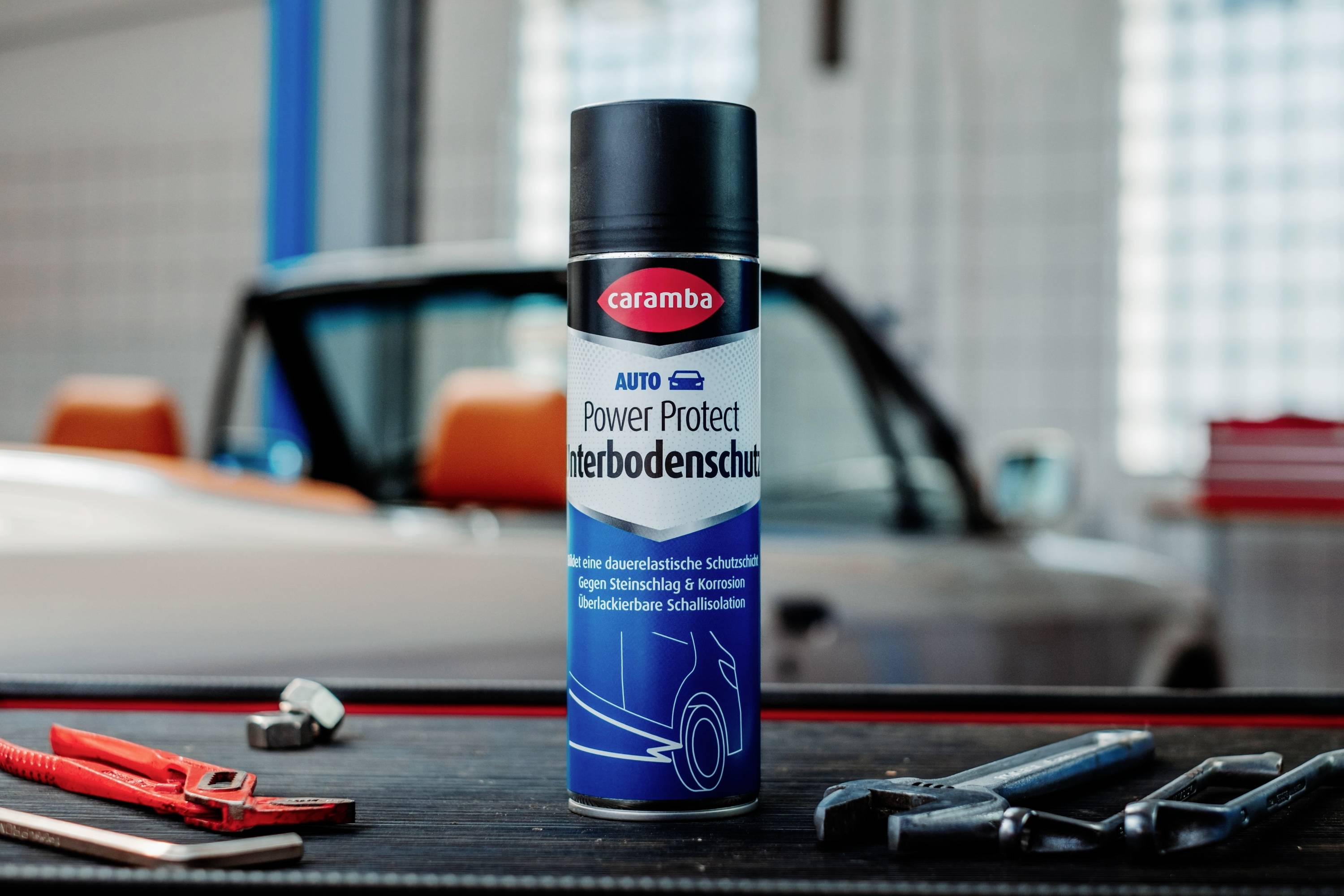 Eine Dose 'Caramba Power Protect Unterbodenschutz' Rostschutzmittel steht auf einem Tisch mit Werkzeugen, vor einem Oldtimer.