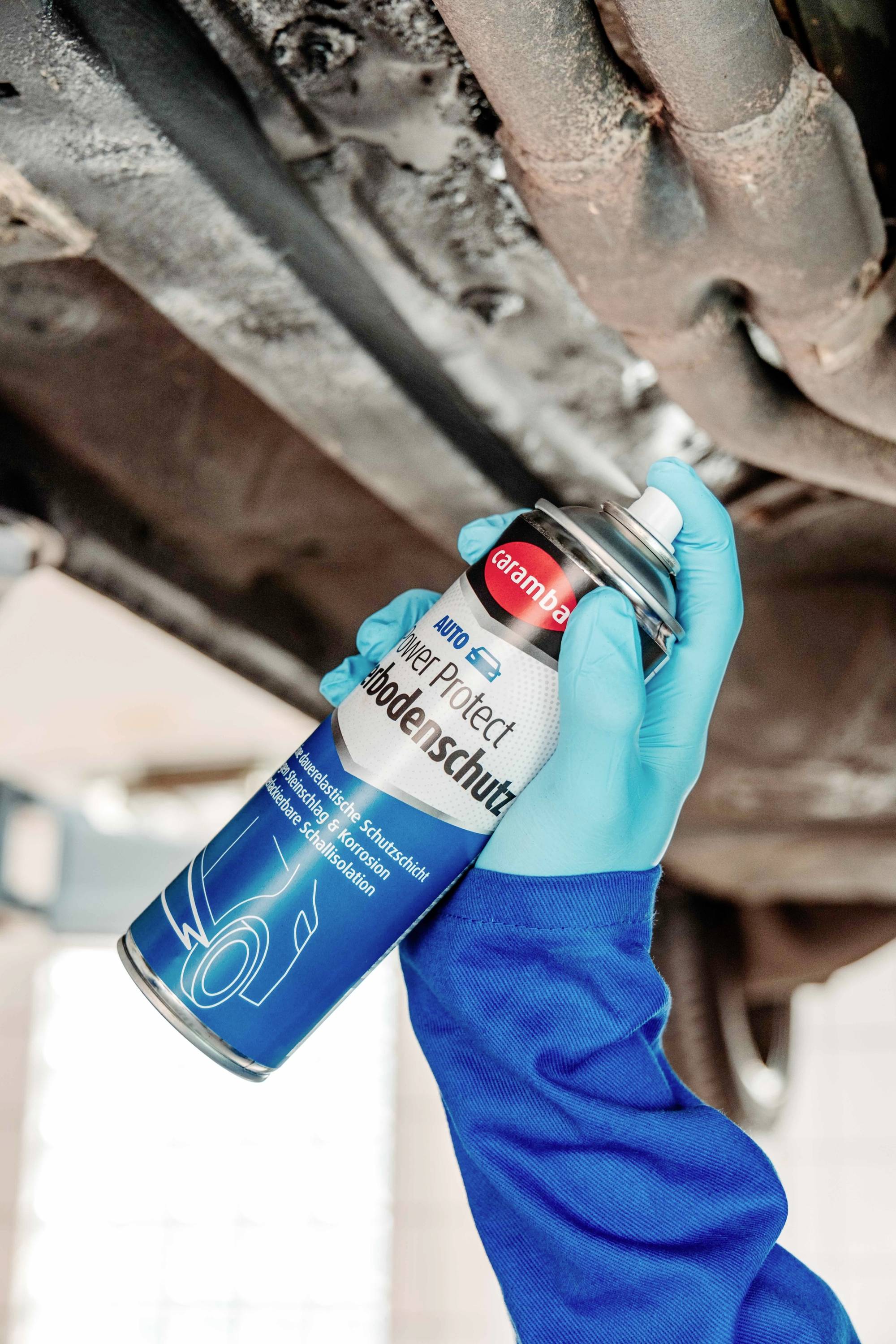 Eine Person mit Handschuhen trägt Unterbodenschutz auf die Unterseite eines Fahrzeugs auf, um Rost und Korrosion zu verhindern und somit die Haltbarkeit zu gewährleisten.