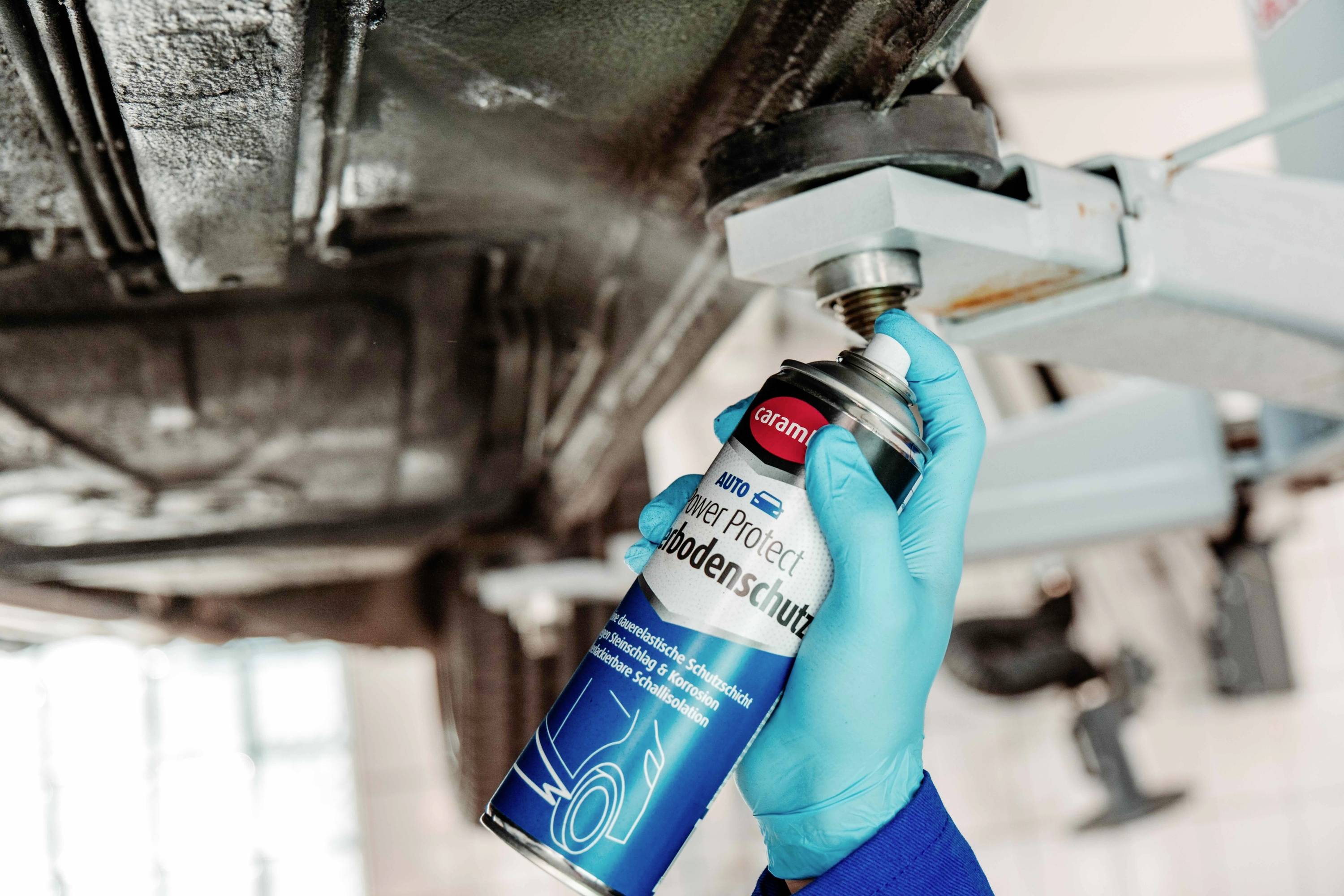 Ein Mechaniker mit blauen Handschuhen trägt Schutzspray auf den Unterbodenbereich eines Autos mit einer Dose mit der Aufschrift „Power Protect Bodenschutz
