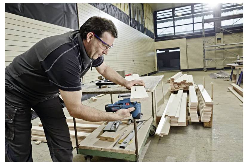 Mann, der einen Elektroschrauber verwendet, um Holzbretter in einer Werkstatt zusammenzubauen, mit Holz, das auf einer Werkbank und dem Boden in der Nähe gestapelt ist.